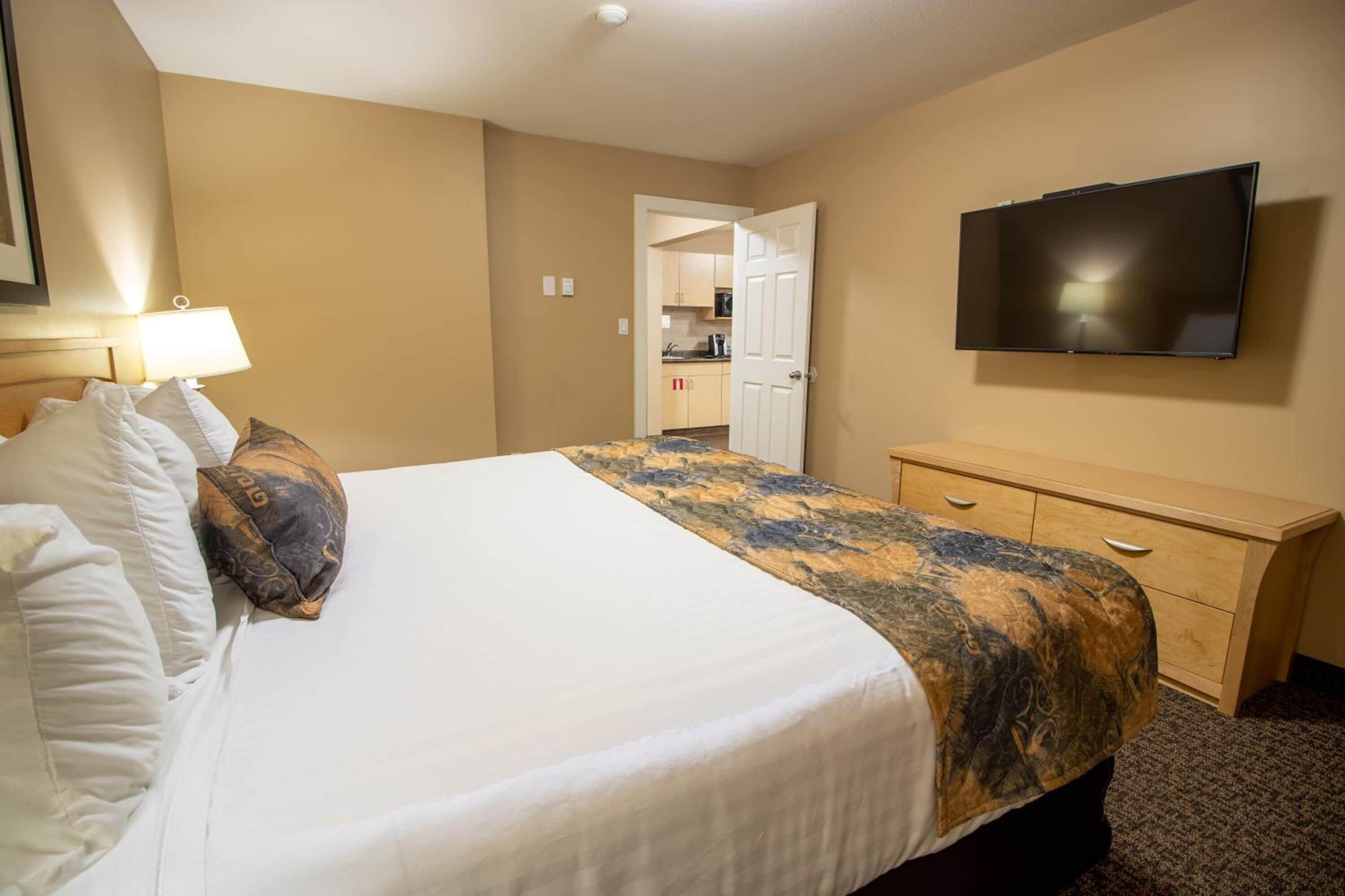 A hotel room with a large bed, a TV mounted on the wall, a desk, and a door leading to a kitchen area.