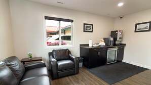 Una sala moderna con un sofá de cuero, un sillón de cuero negro, una mesita con una revista, una ventana con vistas a un edificio y flores, y una kitchenette con una cafetera y armarios.