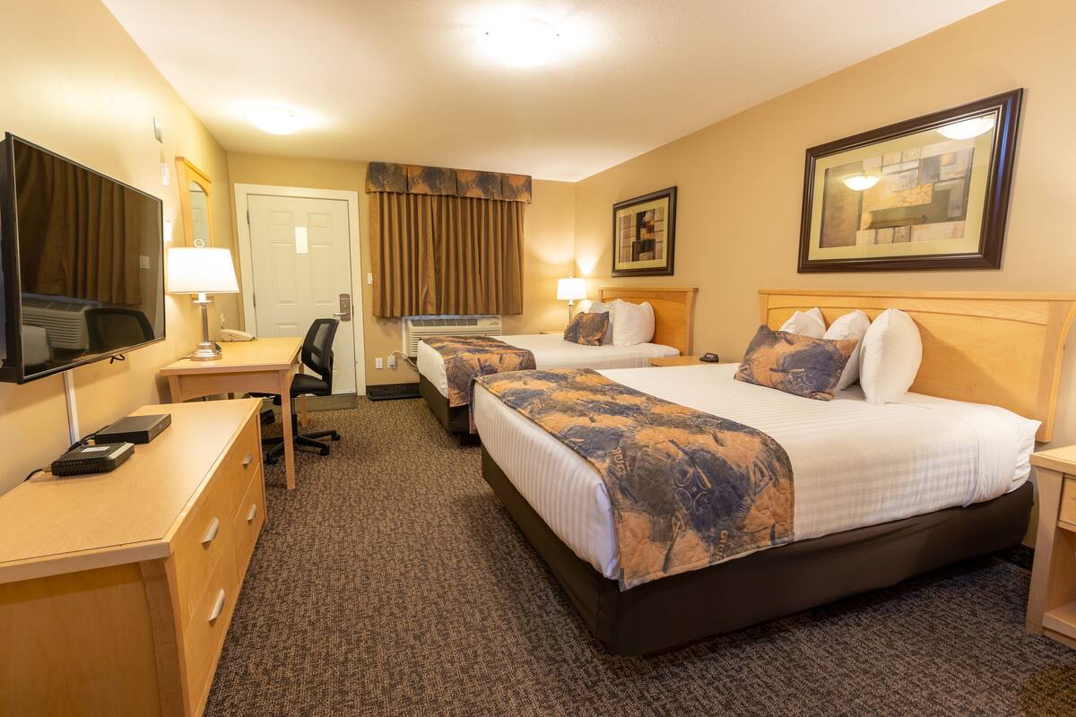 A hotel room with two beds, a desk, a chair, a TV, and a lamp.