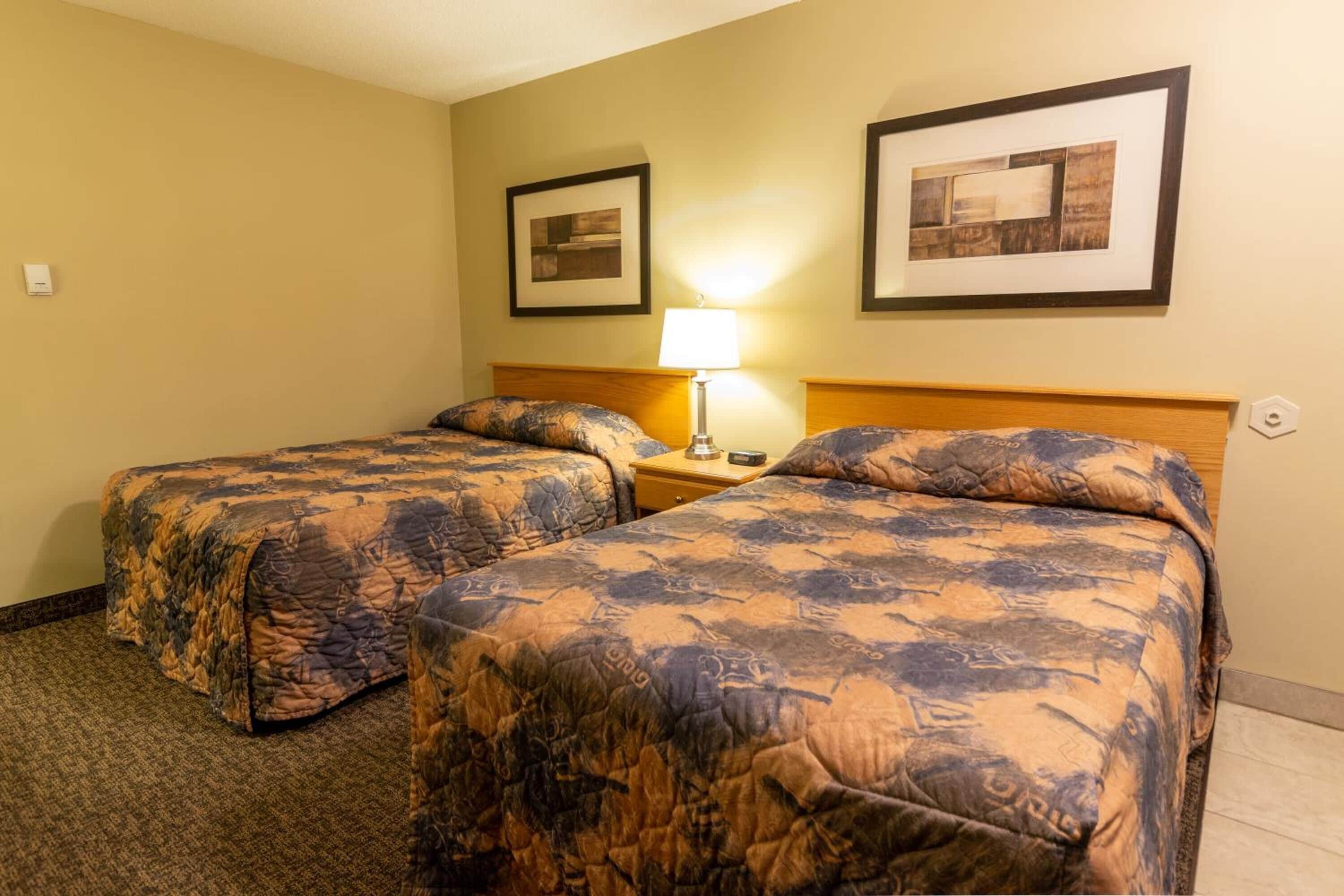 A hotel room with two beds, a nightstand, a lamp, and two framed pictures on the wall.