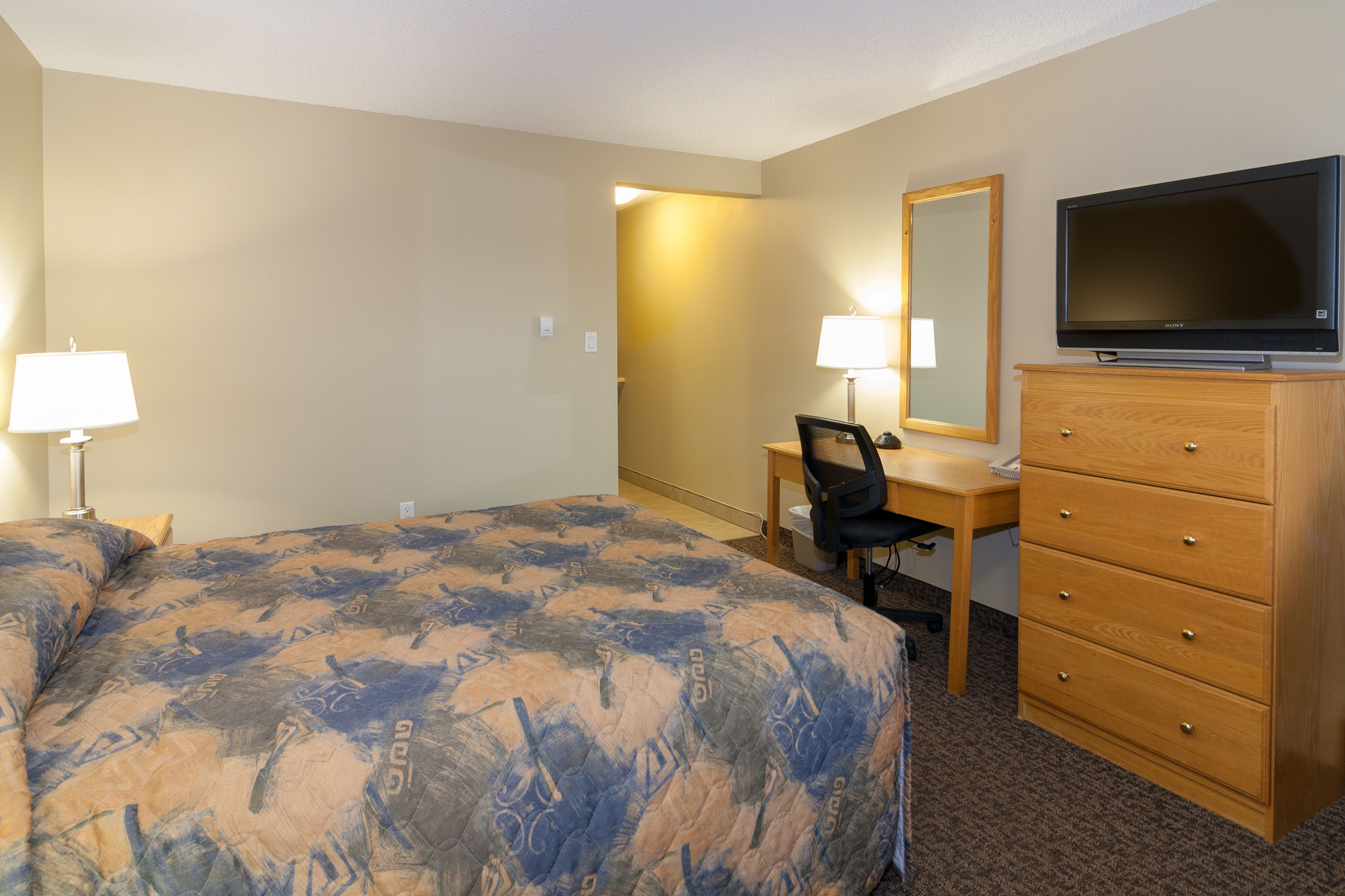 A hotel room with a bed, a desk with a chair, a dresser, a television, and two lamps.