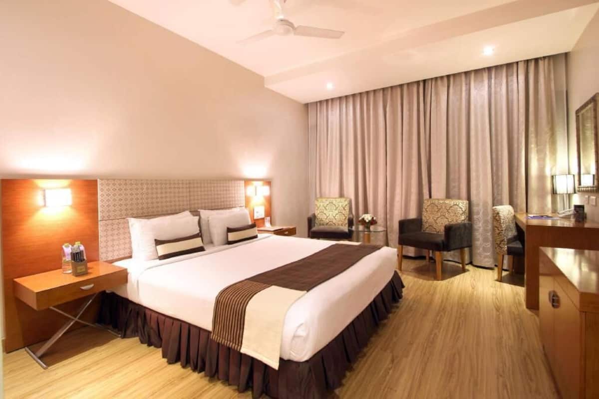 A hotel room with a large bed, two bedside tables, a desk, a chair, a sofa, and a large window with curtains.