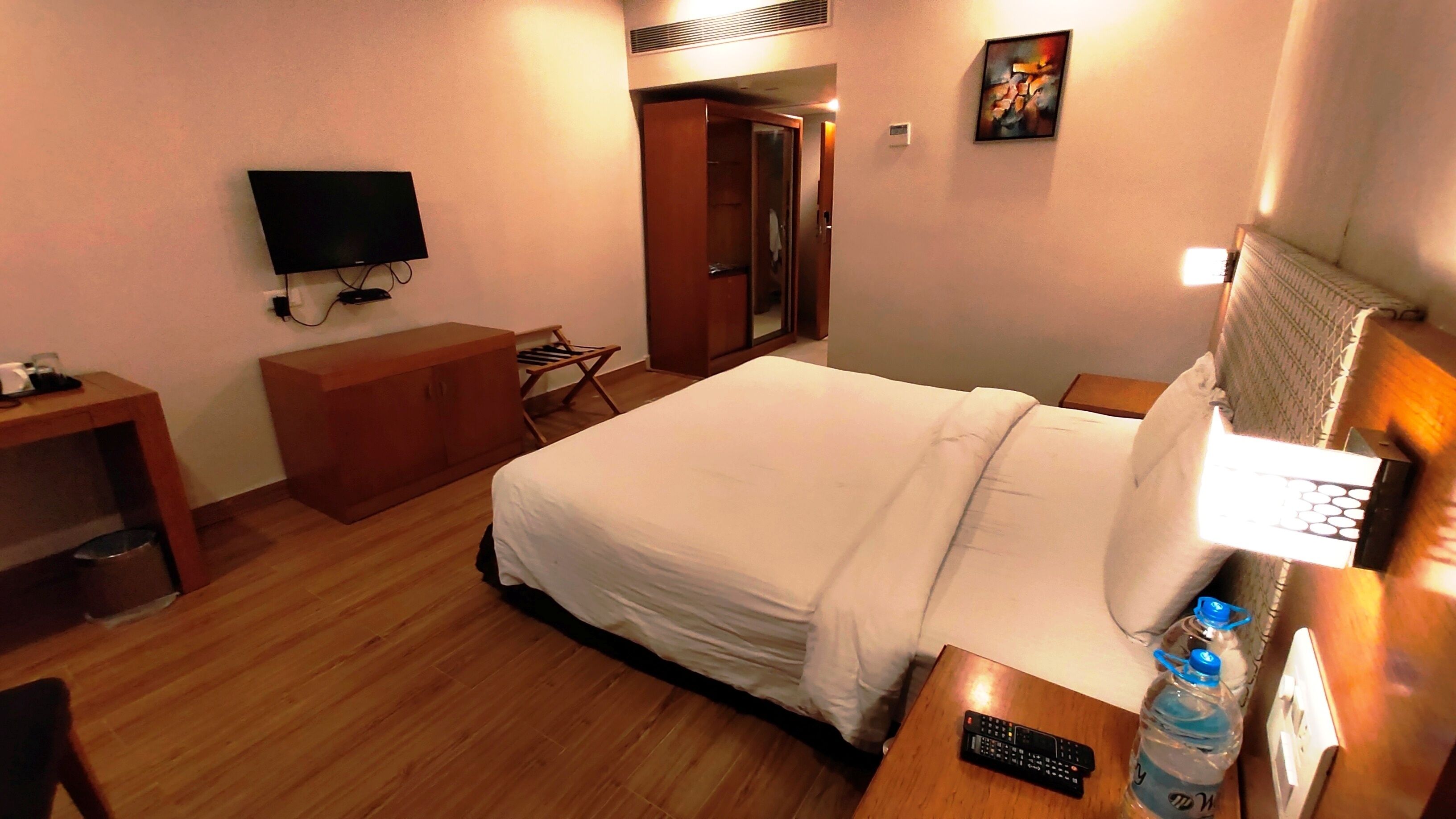 A hotel room with a bed, a TV, a desk, a wardrobe, and a painting on the wall.