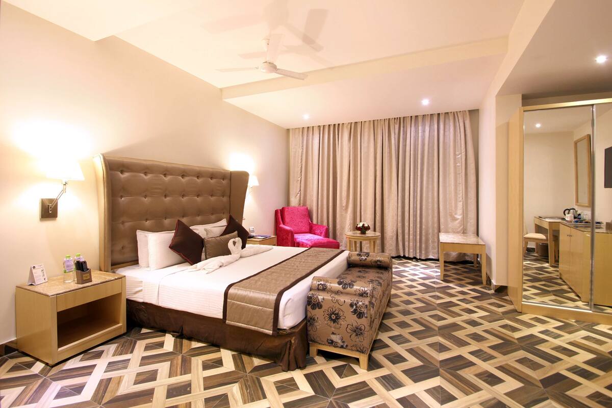 A hotel room with a large bed, a red armchair, a small table, and a mirror.