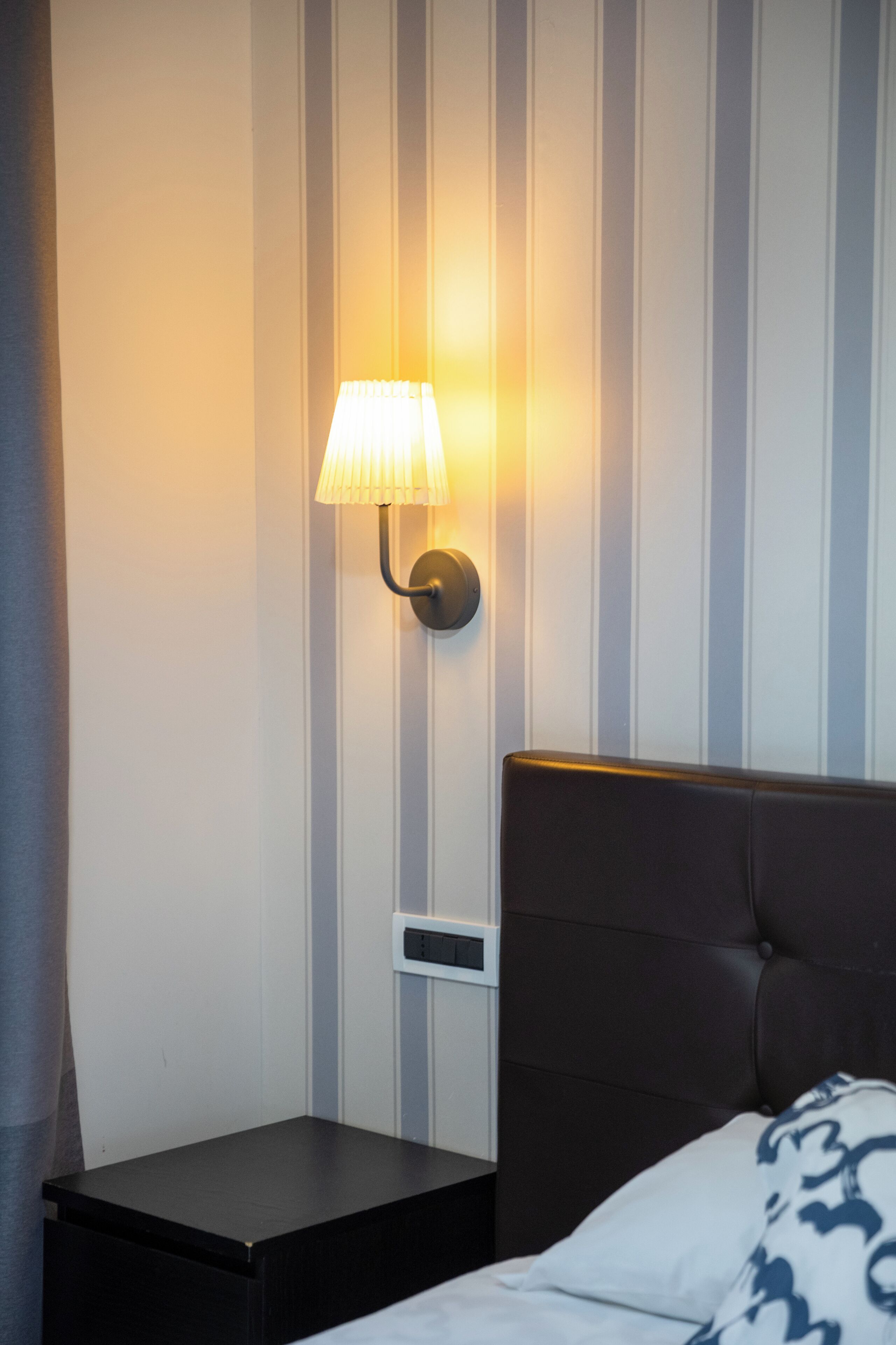 Habitación de hotel con pared a rayas, lámpara de noche, cabecero oscuro y cama con manta estampada.