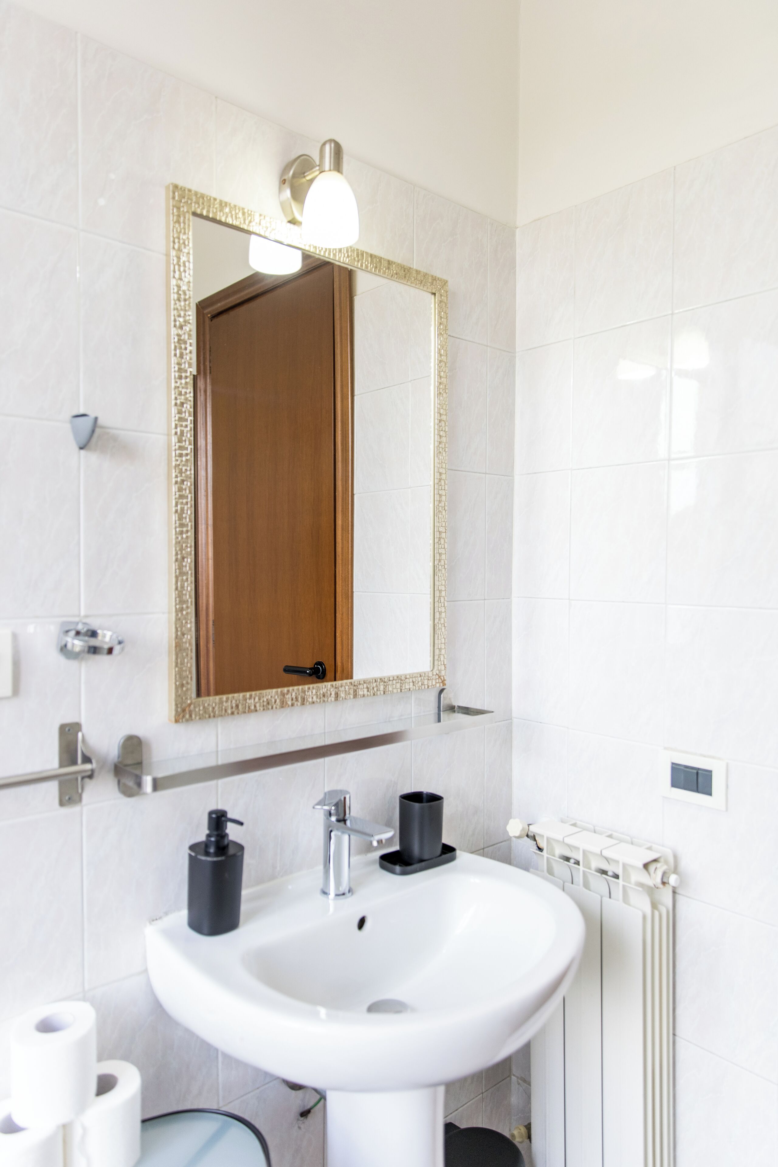 Ein Badezimmer mit einem weißen Waschbecken, einem Spiegel mit goldener Rahmung, einer Holztür und einem weißen Heizkörper.