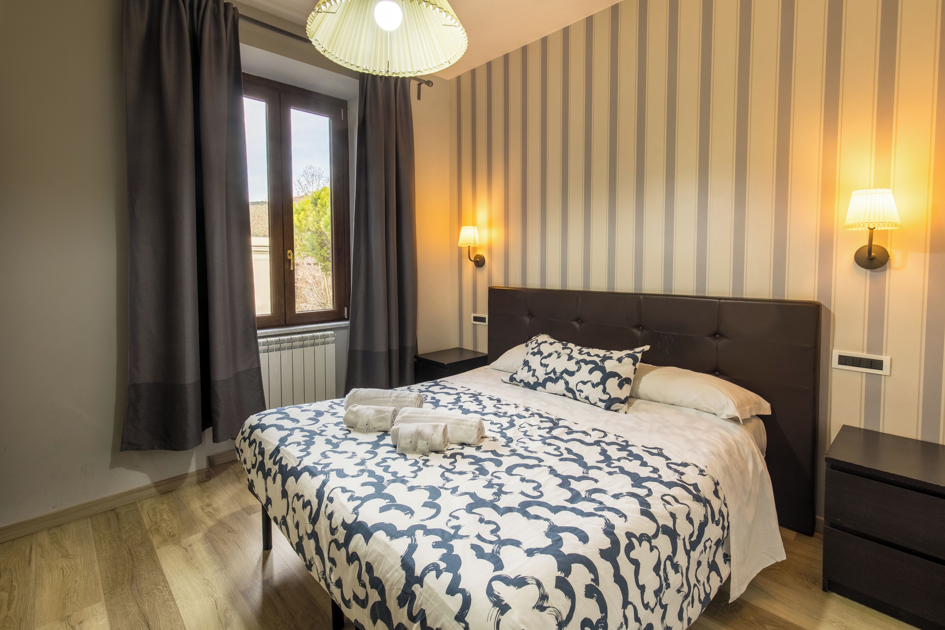 Una habitación de hotel con una cama grande, papel tapiz a rayas, dos lámparas de noche y una ventana con cortinas.