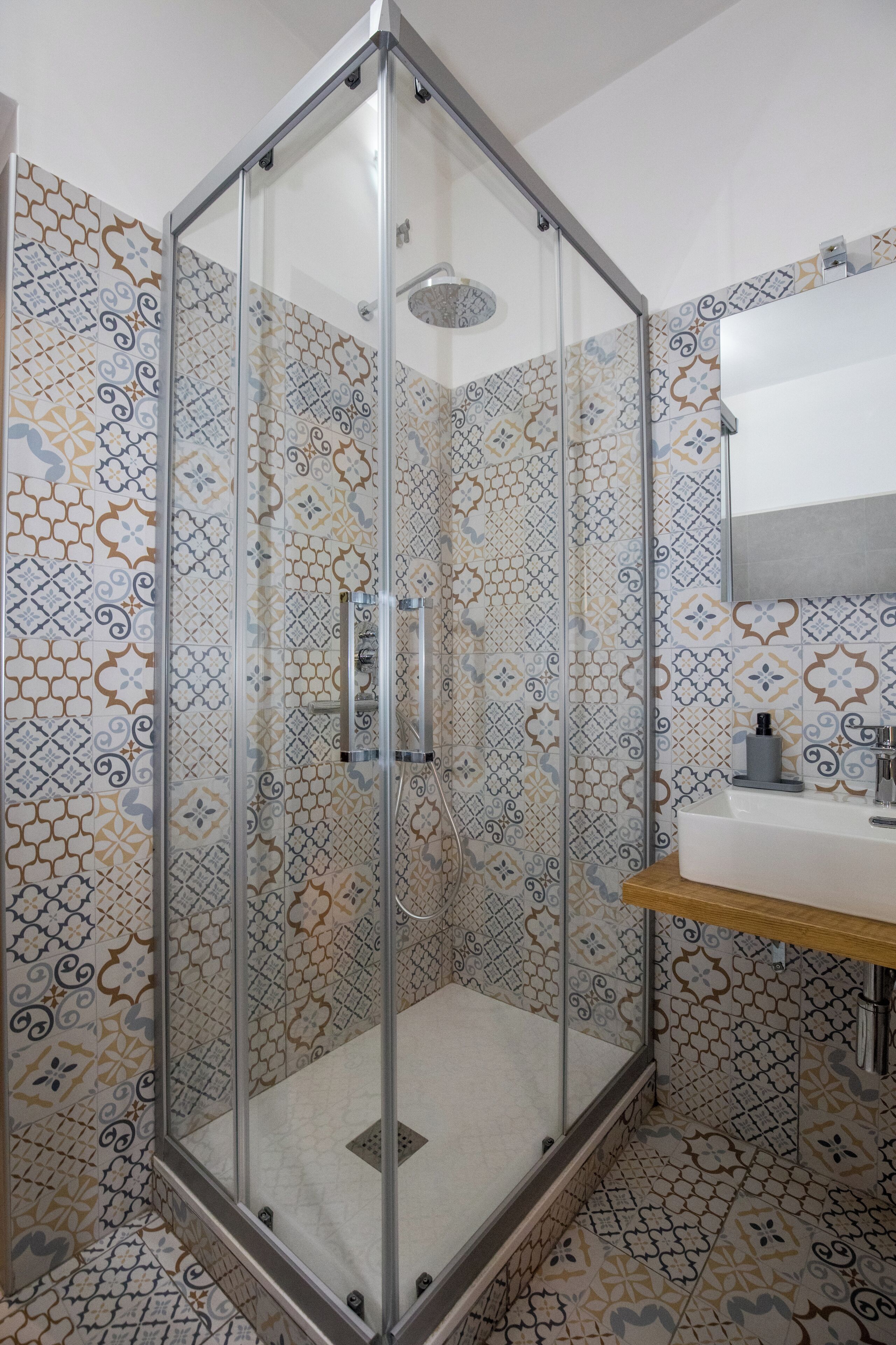 A glass-enclosed shower with patterned tiles, a sink, and a mirror in a bathroom.