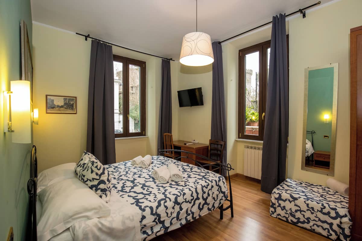A bedroom with a bed, a desk, a chair, a TV, and a window with curtains.