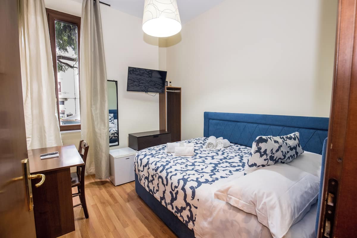 A bedroom with a bed, a desk, a chair, a TV, and a window with curtains.