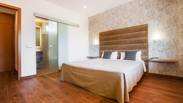 A bedroom with a wooden headboard, a large bed, and a sliding door leading to a bathroom. - Hotel Los Olivos (Getafe)