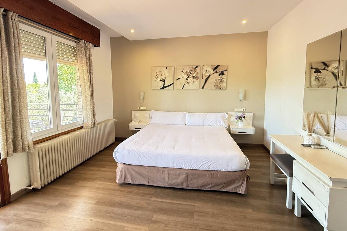 Un dormitorio con una cama grande, un escritorio y una silla. Hay tres lámparas de pared y una ventana con cortinas.
