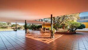 An outdoor covered patio area with tiled flooring, potted plants, and a yellow building in the background. - Hotel Los Olivos (Getafe)