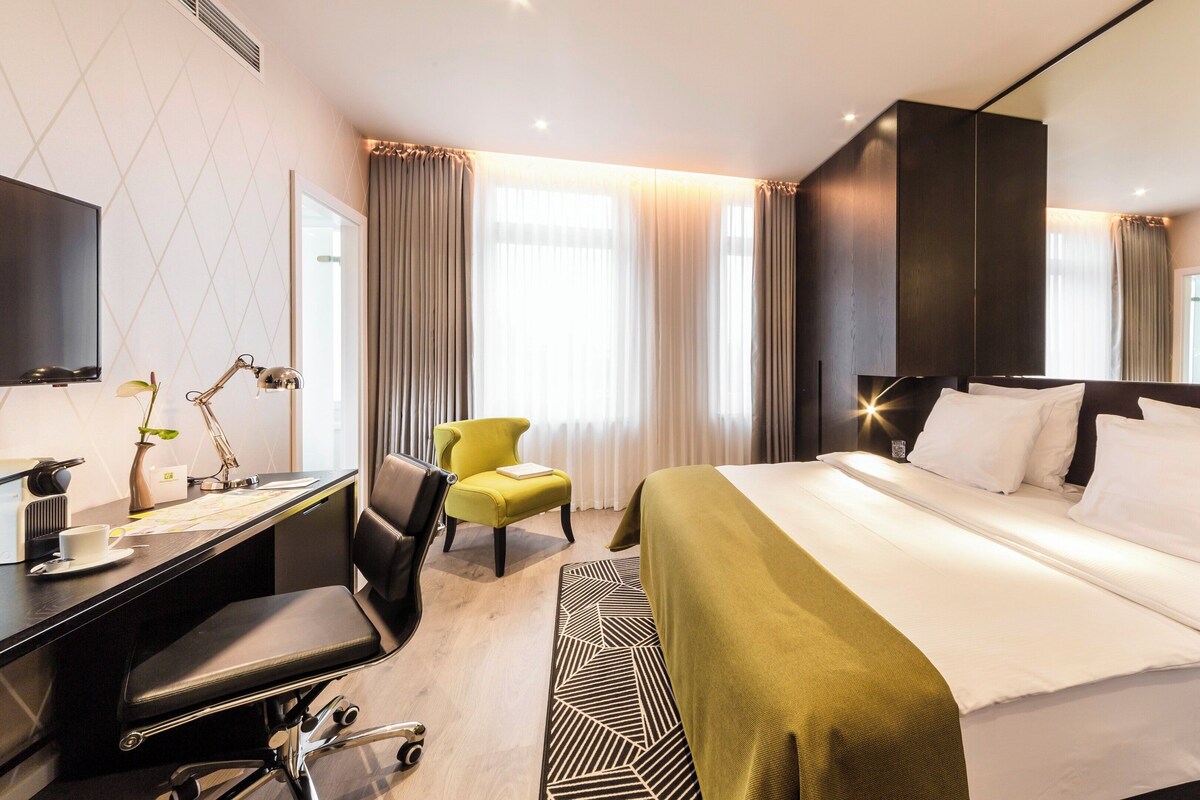 Ein Hotelzimmer mit Bett, Schreibtisch, Stuhl und einem großen Fenster mit Vorhängen.