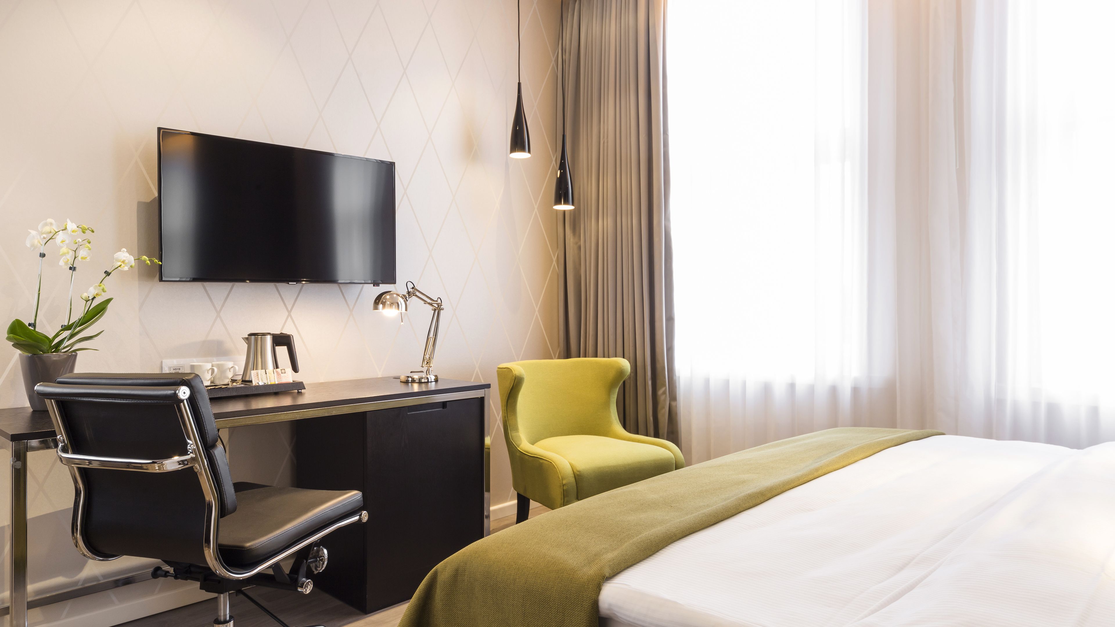 A hotel room with a bed, a desk with a chair, a television, a lamp, and a window with curtains.