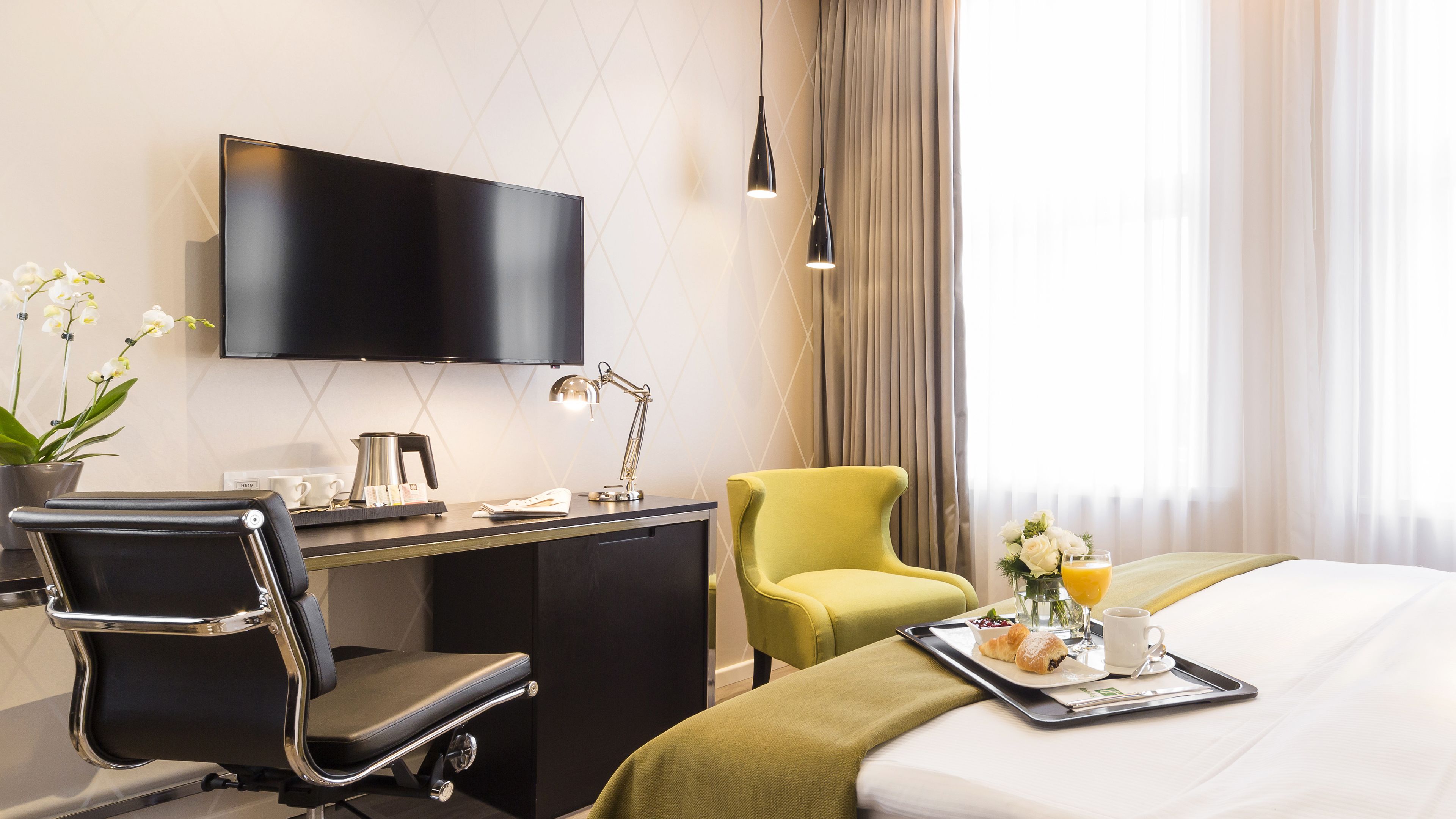 A hotel room with a bed, a desk with a chair, a television, a lamp, a tray with breakfast items, and a window with curtains.