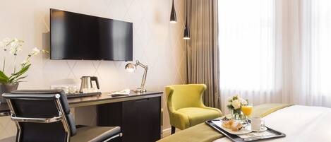 A hotel room with a bed, a desk with a chair, a television, a lamp, a tray with breakfast items, and a window with curtains.