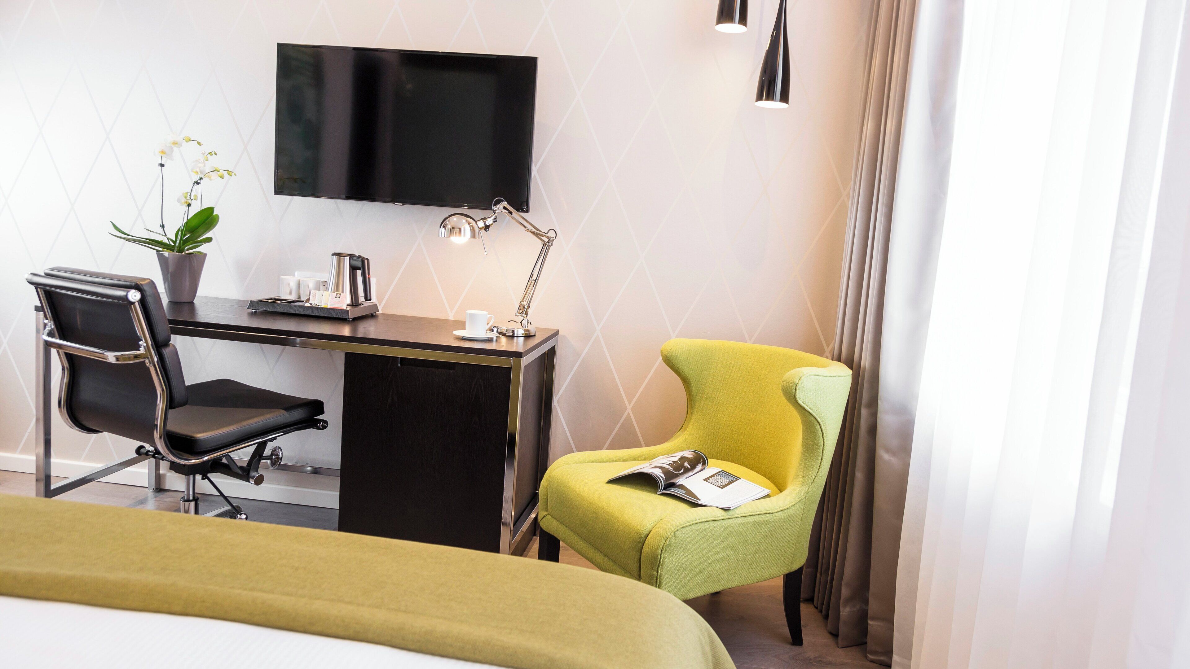A hotel room with a bed, a desk with a chair, a television, a lamp, a potted plant, and a window with curtains.