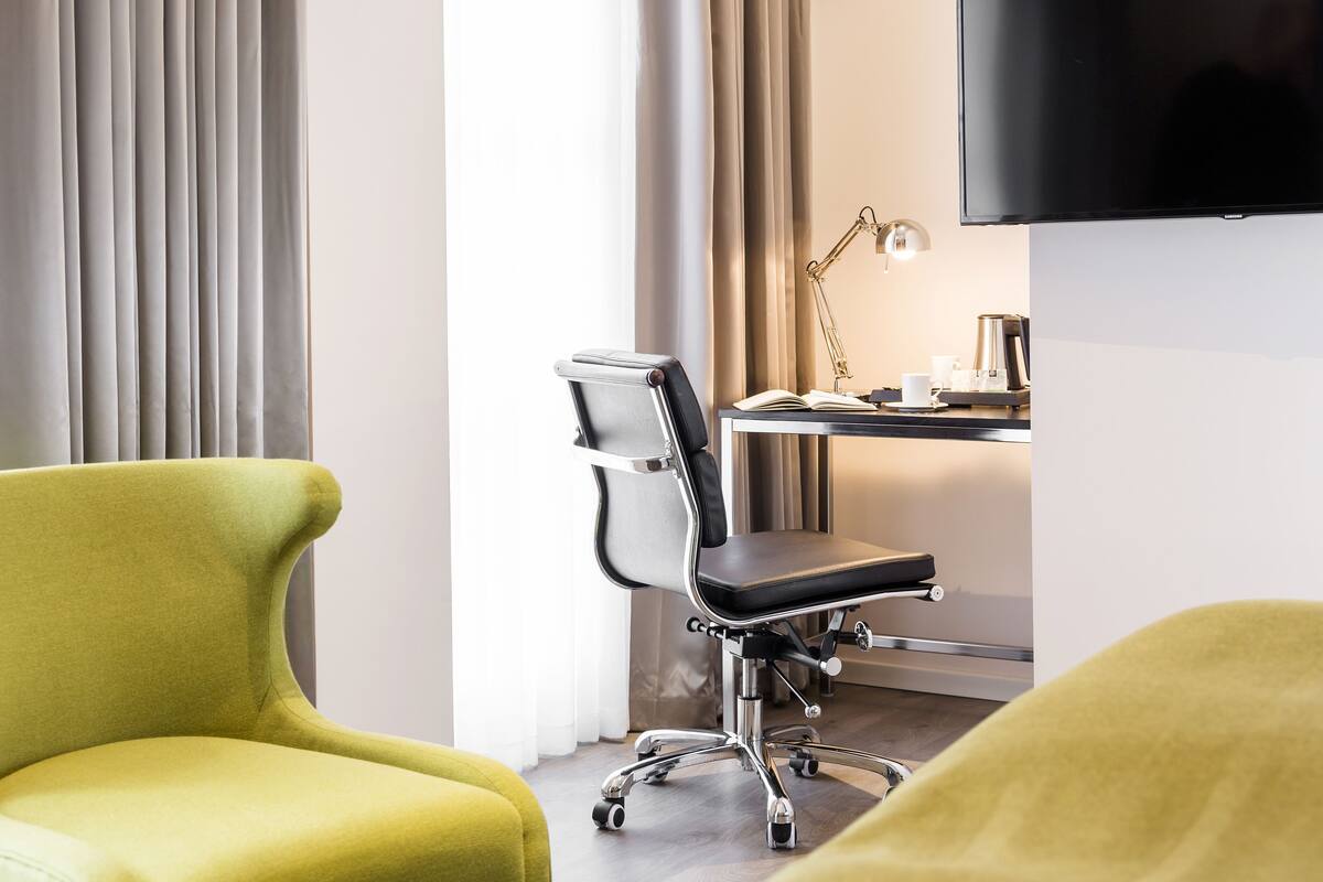 A modern hotel room with a yellow armchair, a black office chair, a desk with a lamp, and a flat-screen TV.