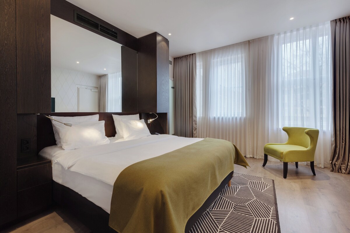 A hotel room with a large bed, a chair, a mirror, and a window with curtains.
