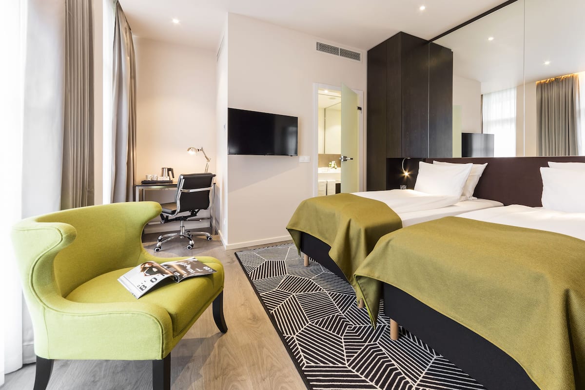 A hotel room with two beds, a desk with a chair, a TV, and a green armchair.