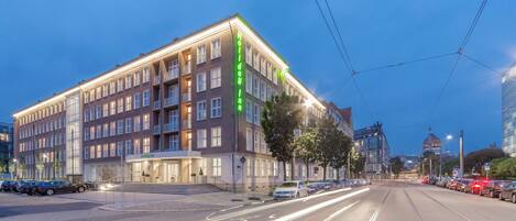 A multi-story hotel with a prominent 'Holiday Inn' sign, a tram line nearby, and a parking lot with cars.
