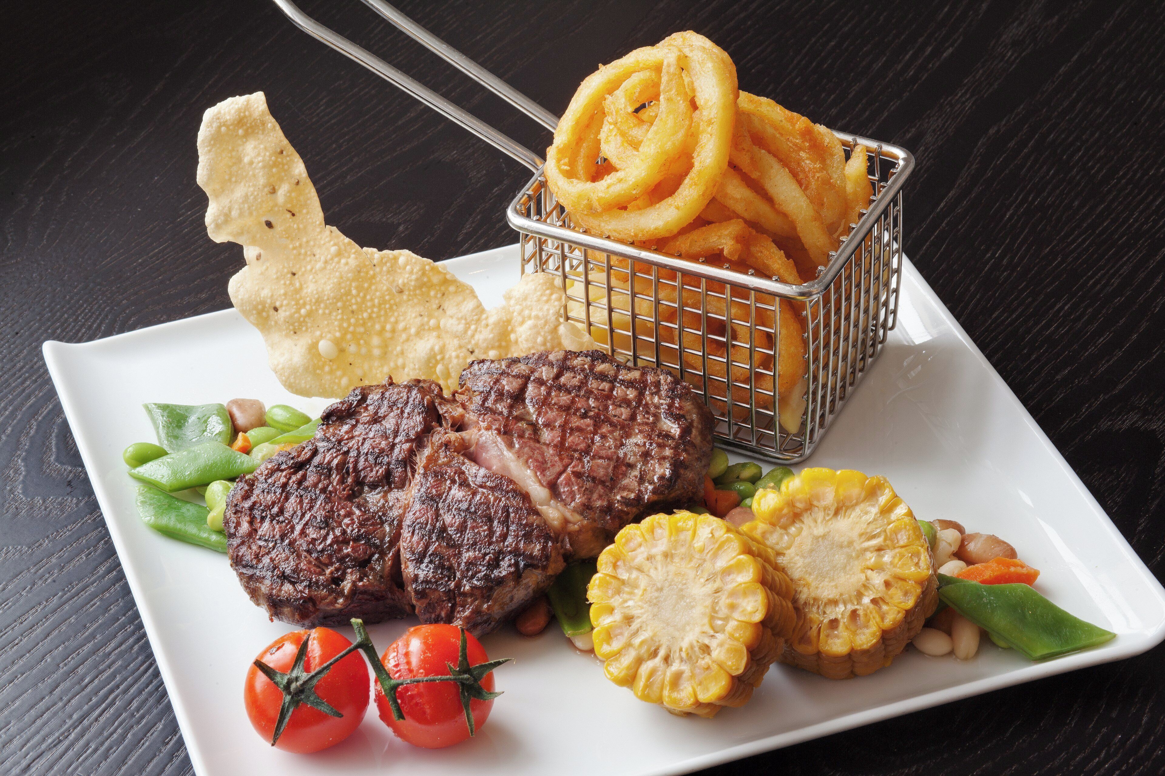 Ein Teller mit einem Steak, Mais, Tomaten und einer Körbchen voller Zwiebelringe.