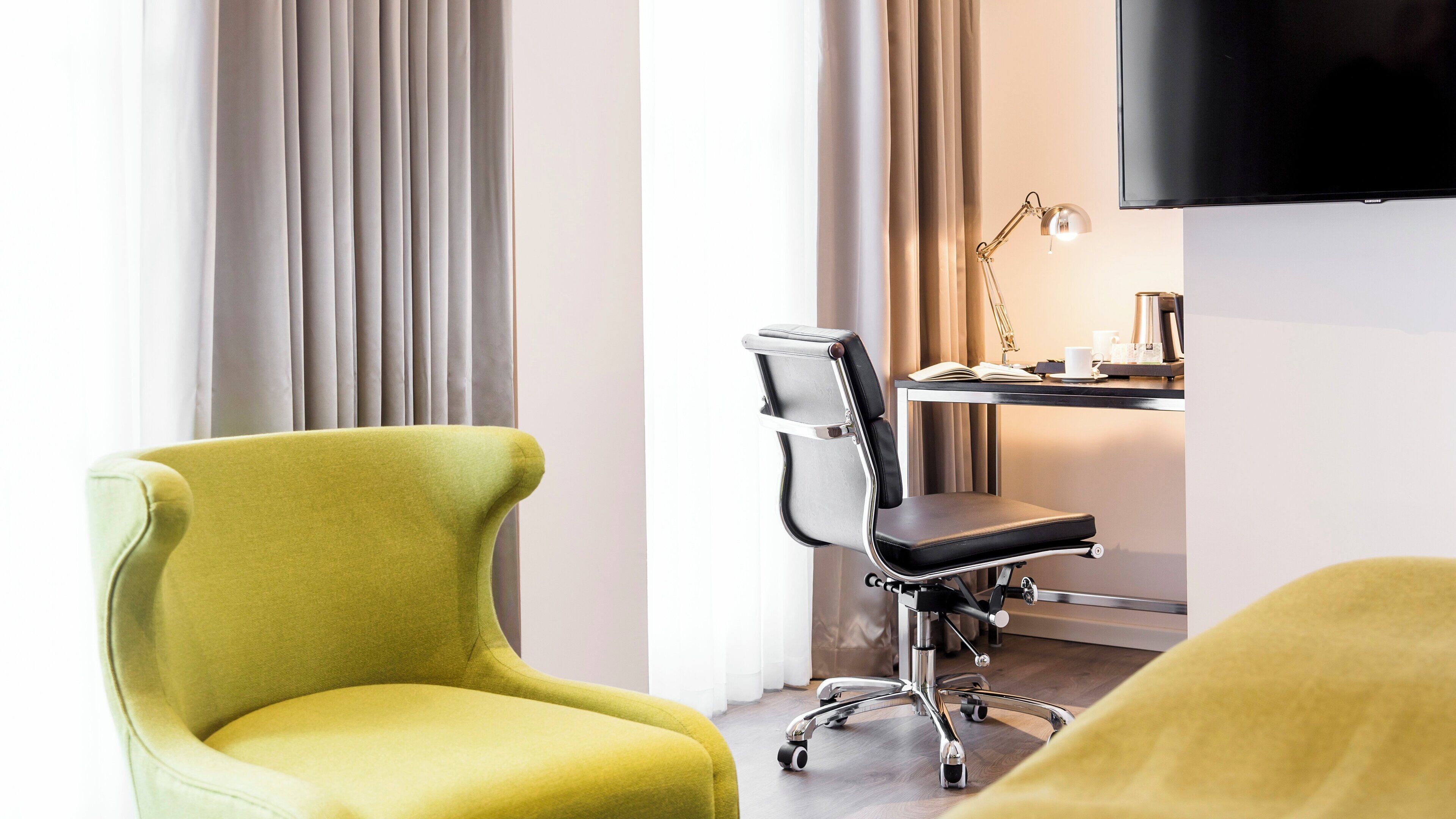 A modern hotel room with a yellow armchair, a black office chair, a desk with a lamp, a coffee maker, and a flat-screen TV.