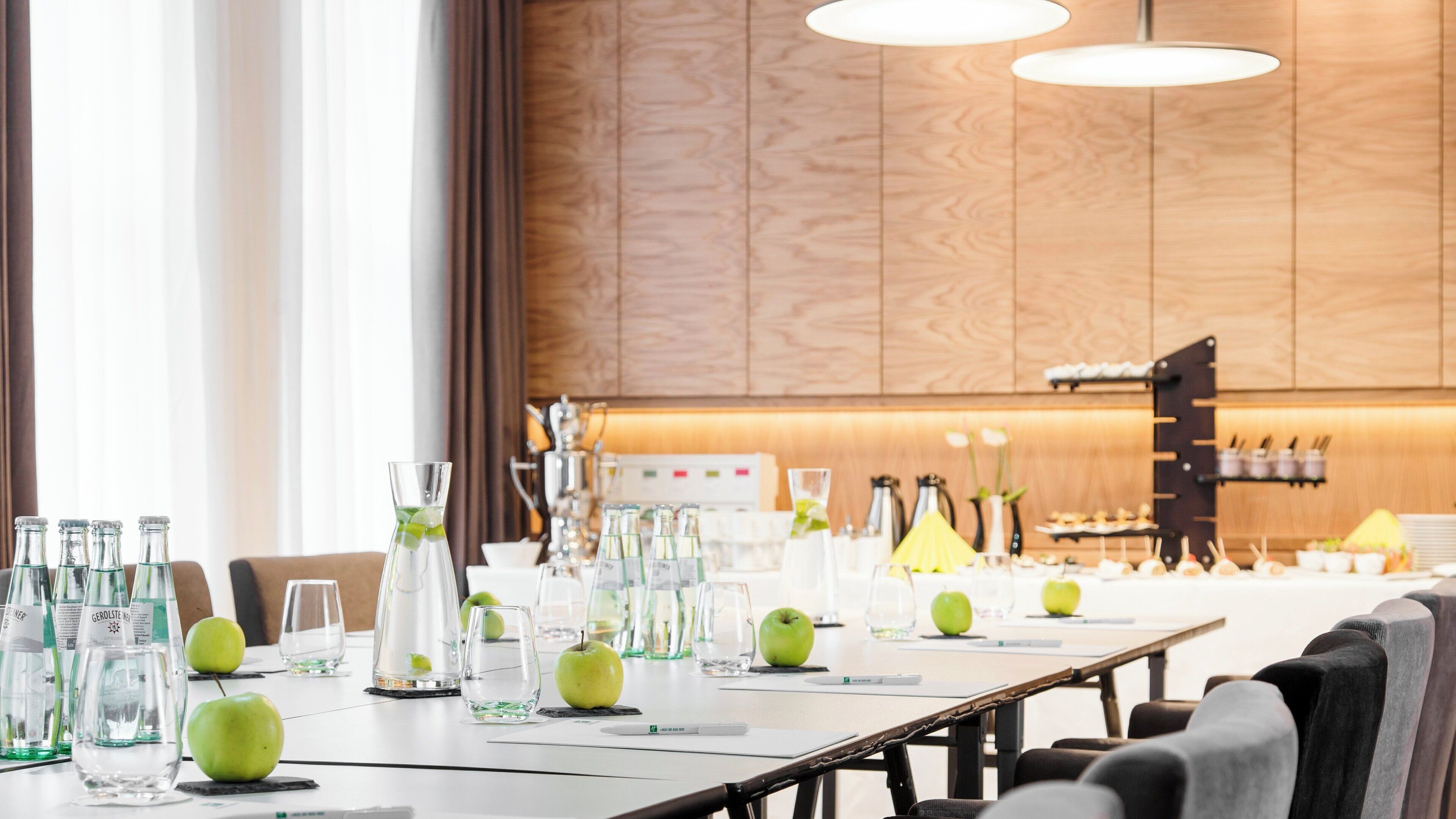 A conference table set with glassware, apples, and a water dispenser in a modern hotel room.
