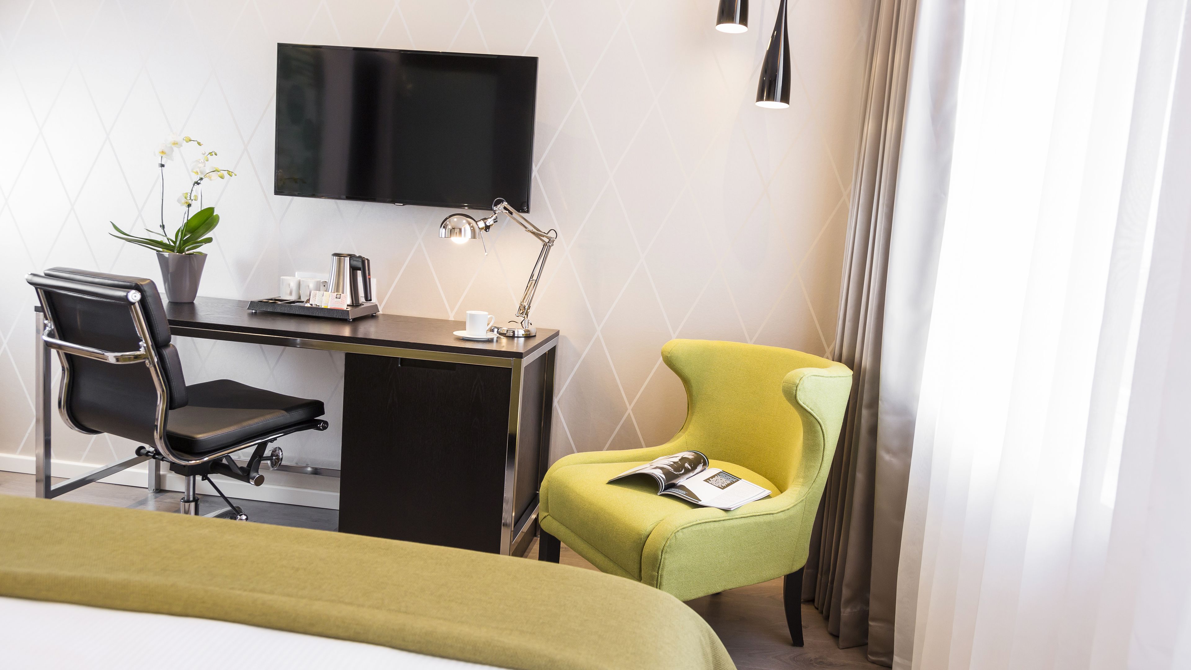 A hotel room with a bed, a desk with a chair, a television, a lamp, and a potted plant.