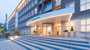 A modern hotel entrance with a curved awning, a revolving door, and a staircase leading up to the entrance. - Holiday Inn Dresden - Am Zwinger by IHG (Dresden)