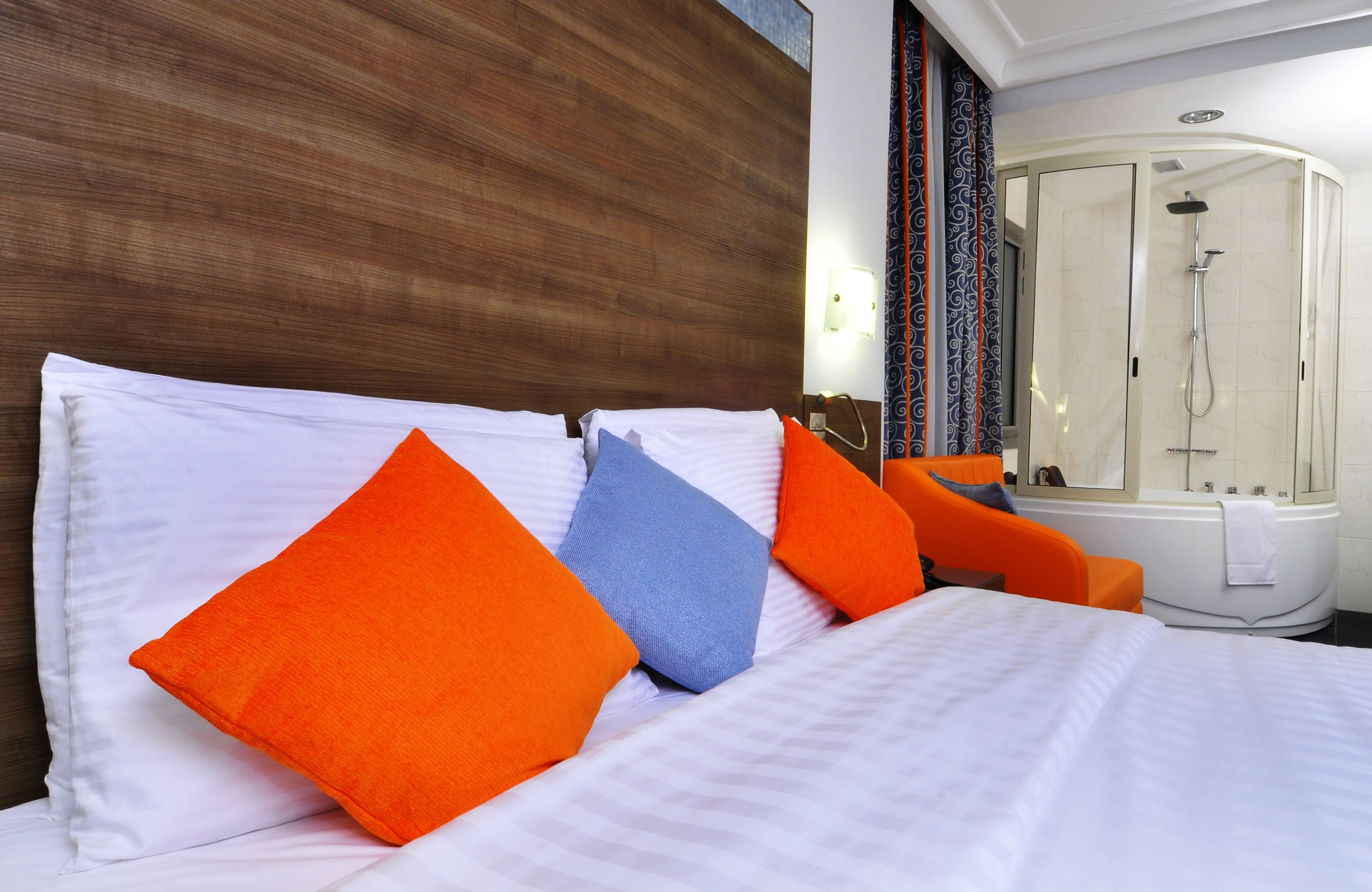 Habitación de hotel con una cama grande, almohadas anaranjadas y azules, cabecera blanca y un baño separado con ducha y bañera.