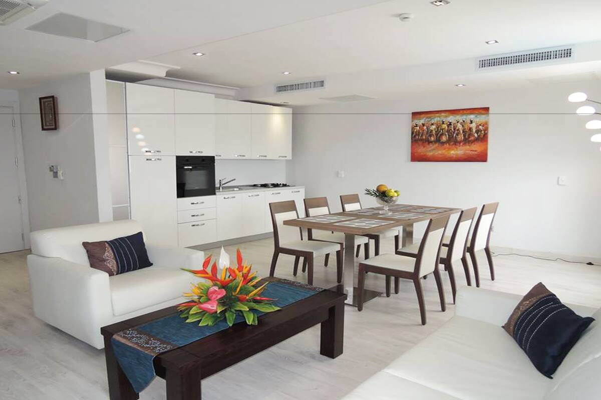 A modern living room with a dining area, white furniture, and a kitchen in the background.