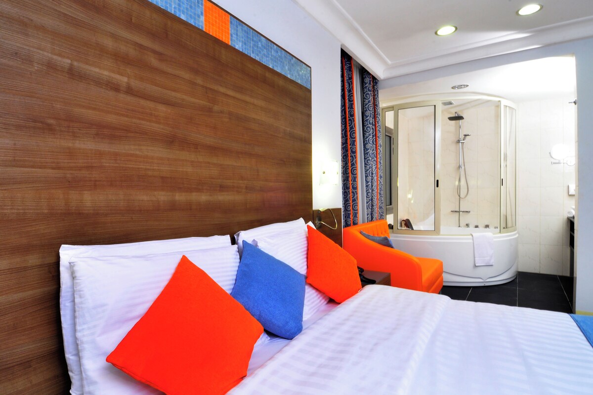 A hotel room with a wooden headboard, a bed with white and orange bedding, a shower area with a glass enclosure, and a bathtub.