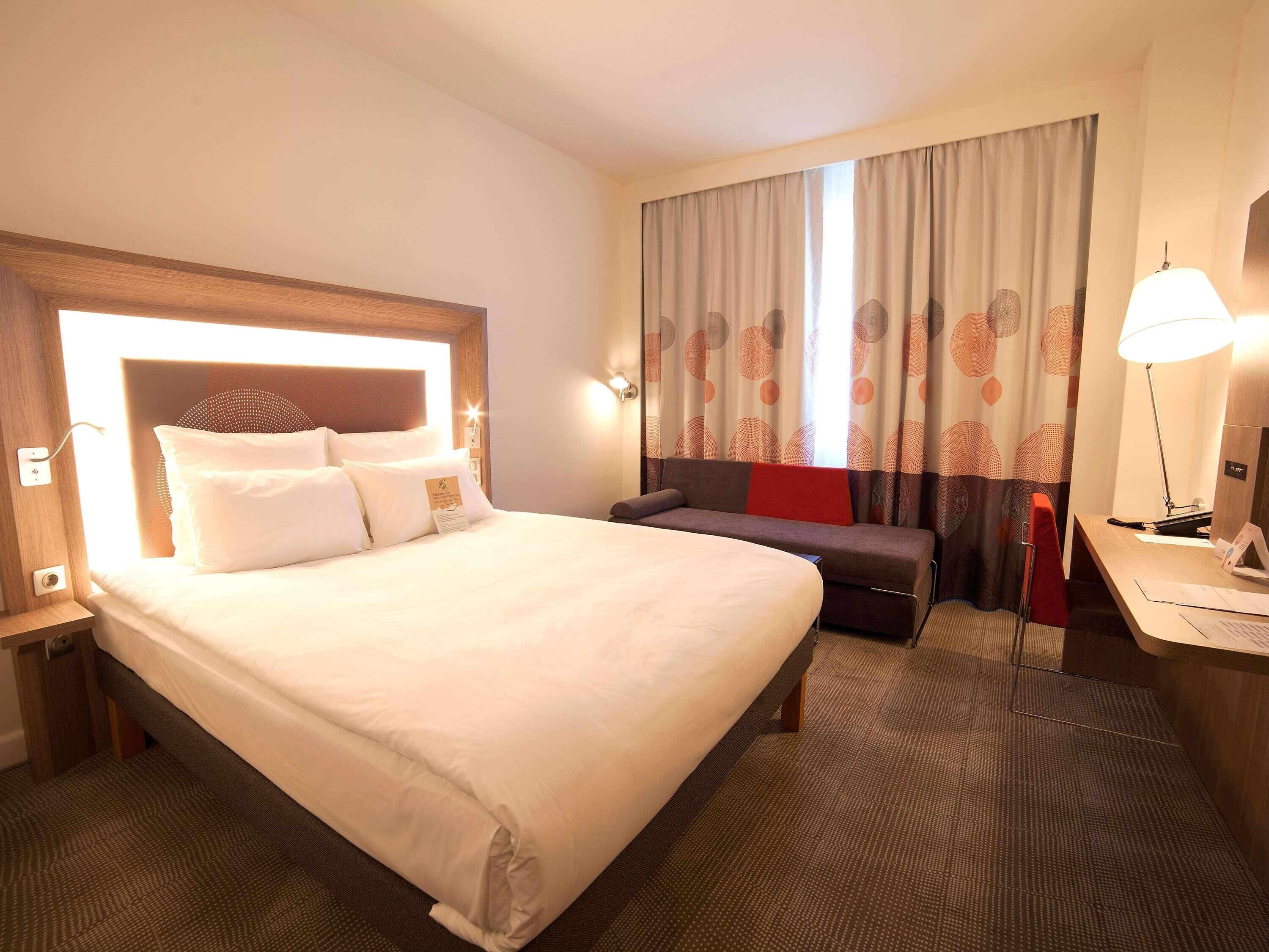 Ein Hotelzimmer mit einem großen Bett, einem Schreibtisch, einem Sessel und einem Fenster mit Vorhängen.