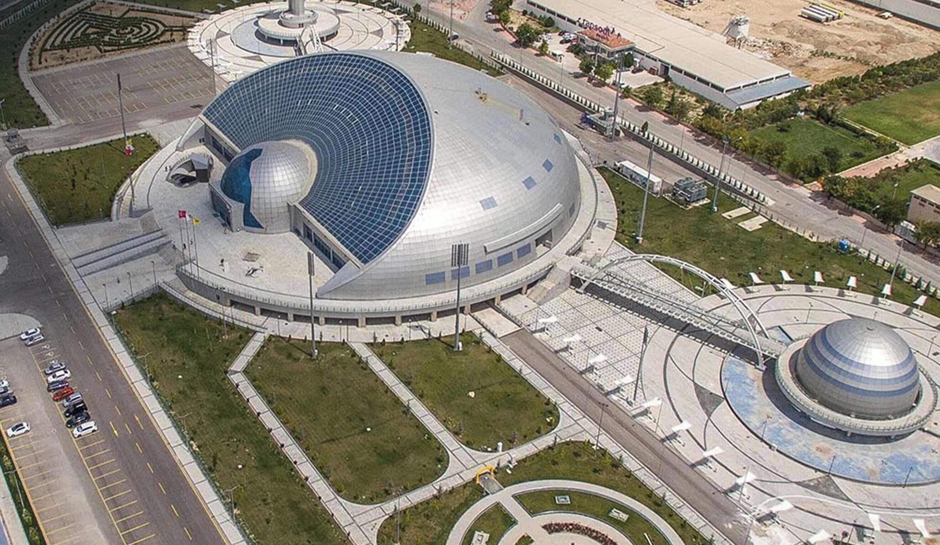 Un grand bâtiment circulaire surmonté d’un dôme, entouré d’un espace paysager agrémenté de sentiers et d’un parking.