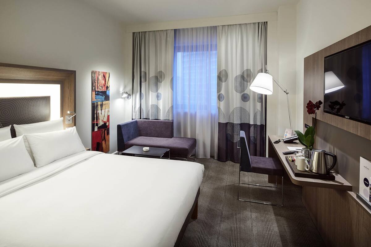 Habitación de hotel con cama, escritorio, silla, televisor, lámpara y ventana con cortinas.
