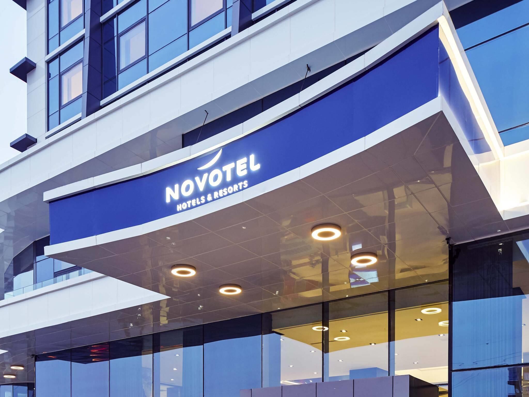 A modern hotel facade with a prominent 'NOVOTEL HOTELS & RESORTS' sign, large windows, and a reflective glass exterior.