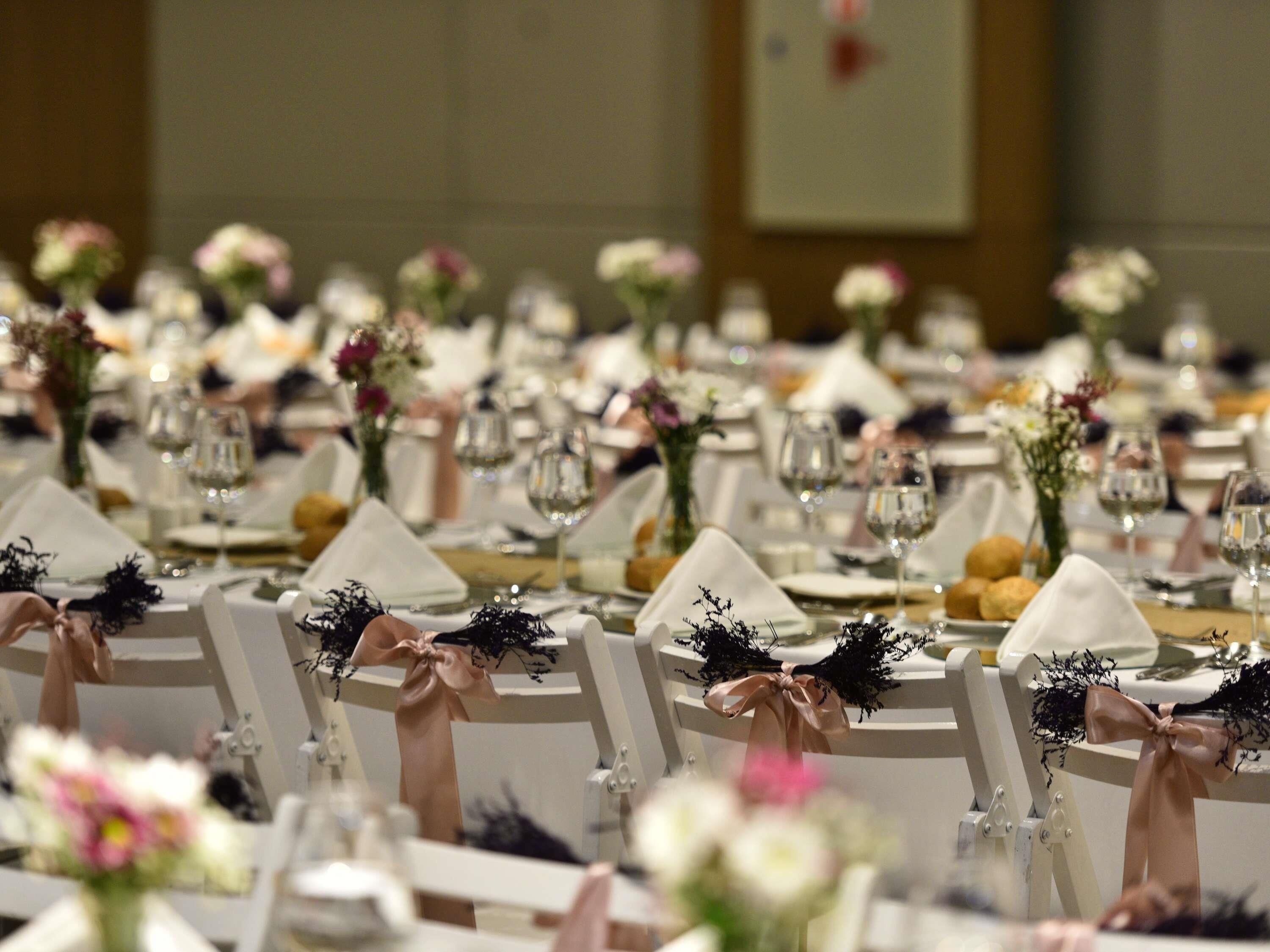 Un salón de banquetes con mesas redondas dispuestas para un evento formal, cada una con mantel blanco, cristalería y centros de mesa florales.