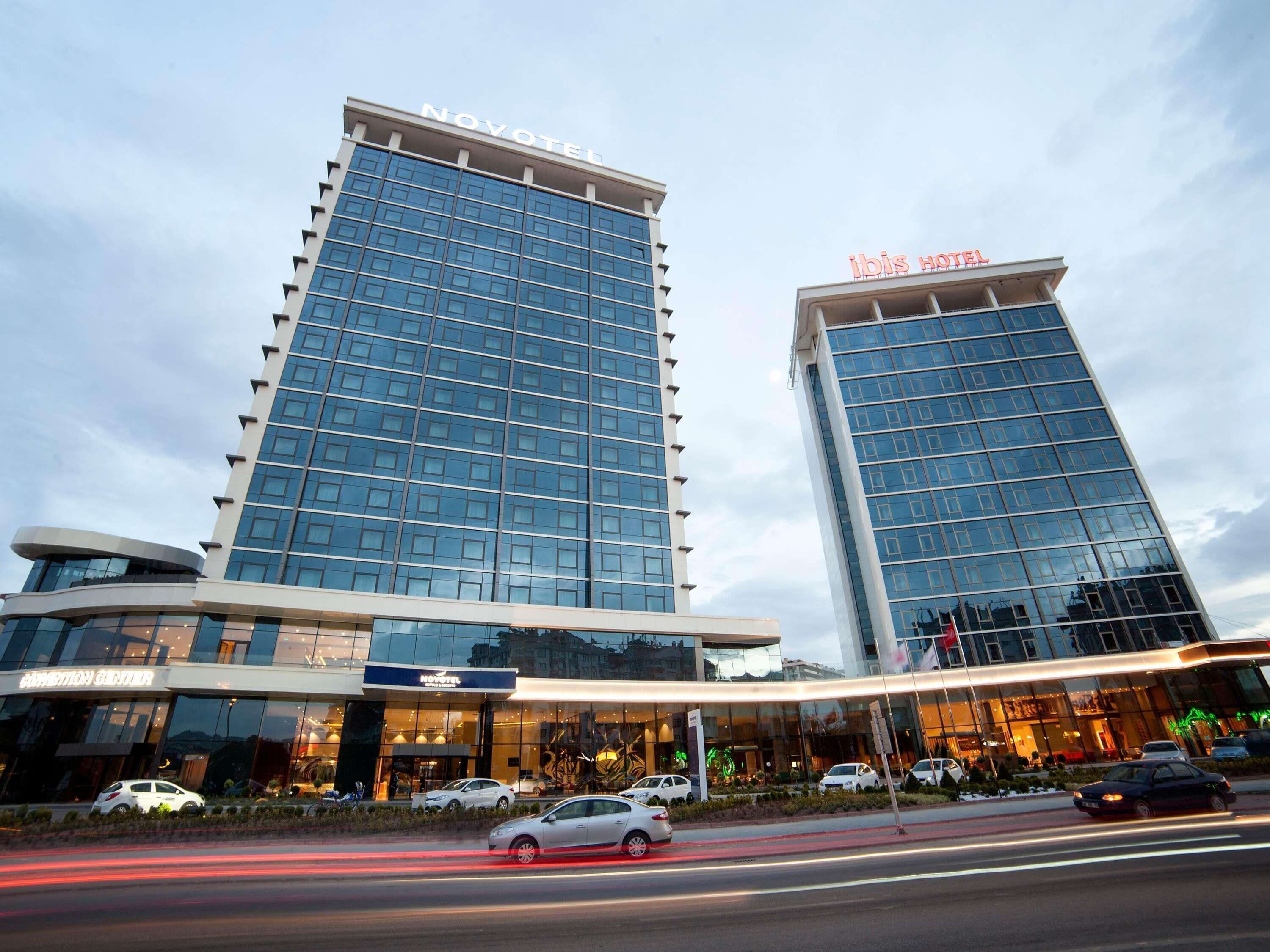 A hotel with a modern glass facade, two visible signage for 'NOVOTEL' and 'IBIS HOTEL', and a parking area with cars.