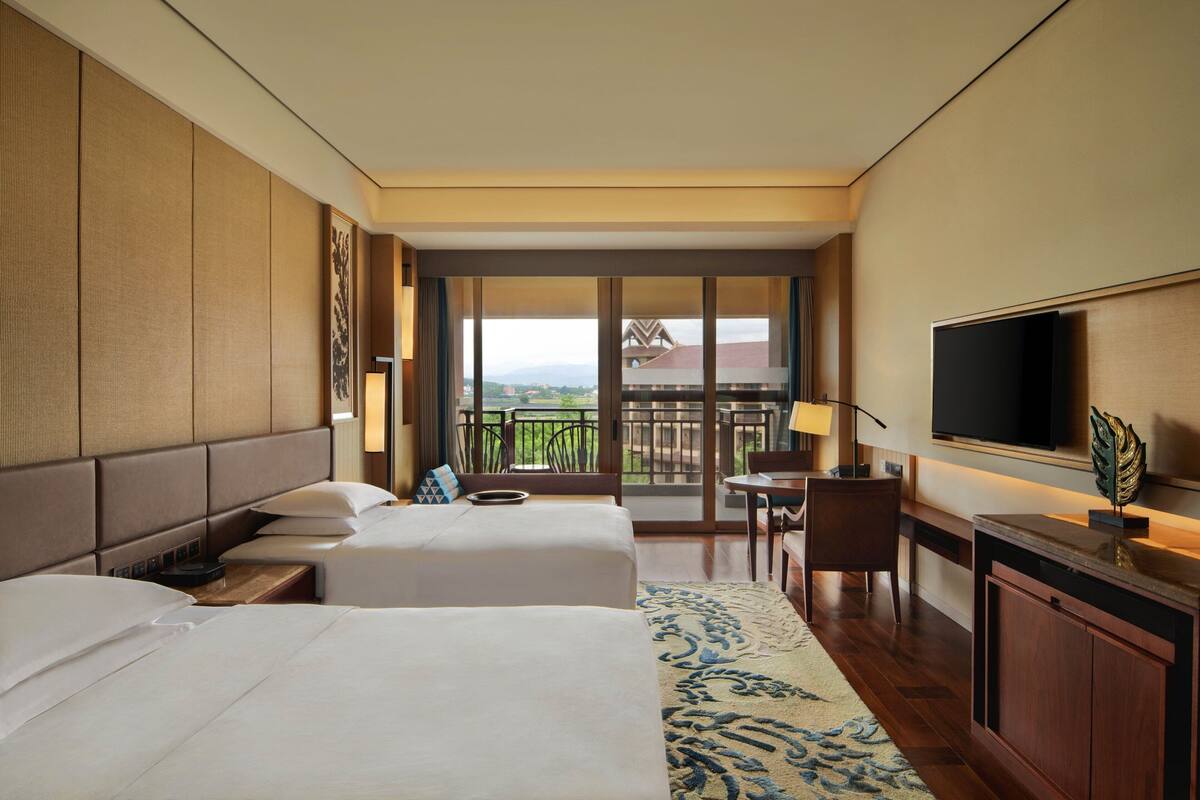 A hotel room with two beds, a TV, a desk, and a balcony with a view.