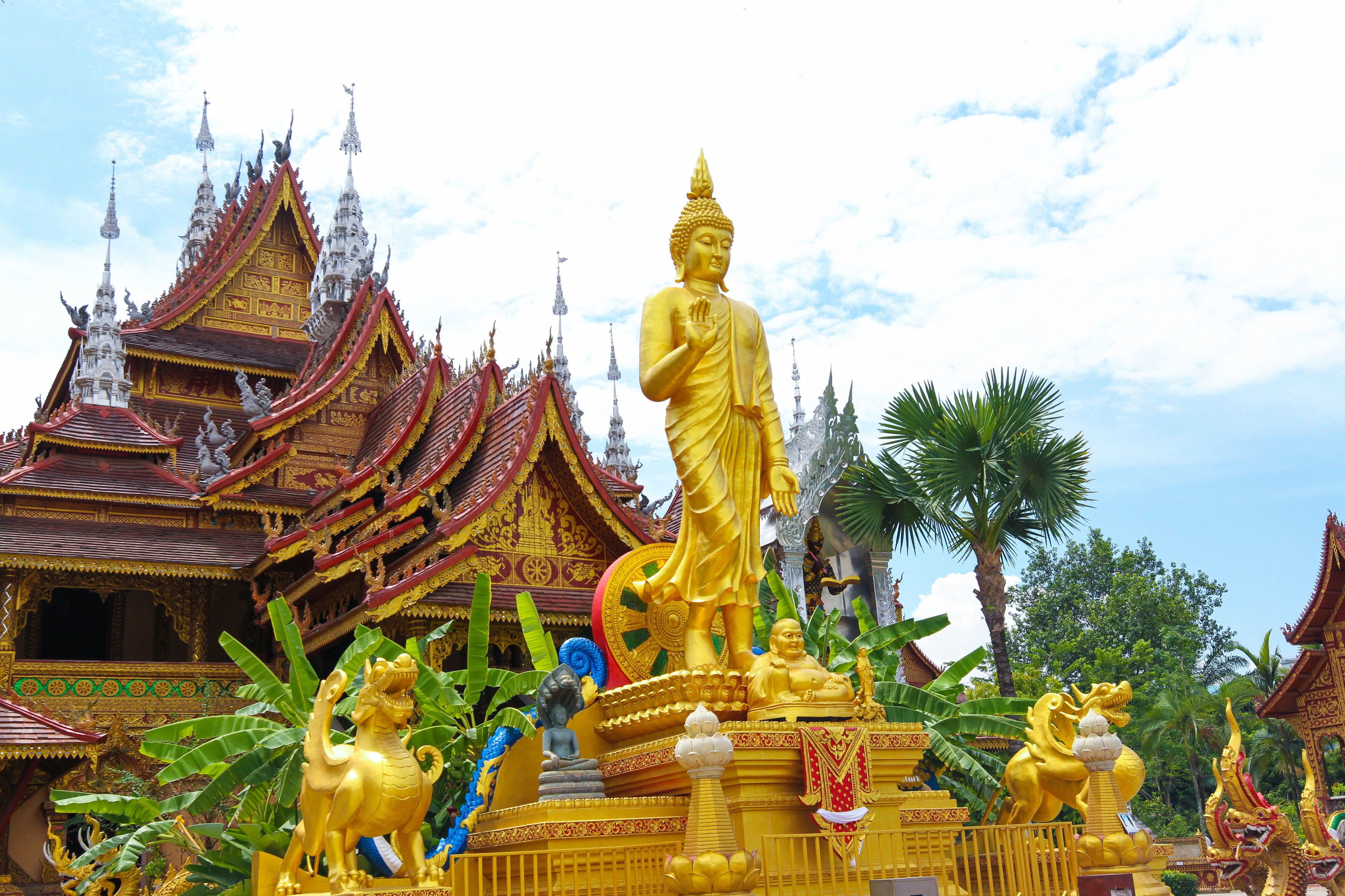 Une grande statue de Bouddha en or se dresse devant un temple thaïlandais traditionnel, dont l’architecture est richement décorée.