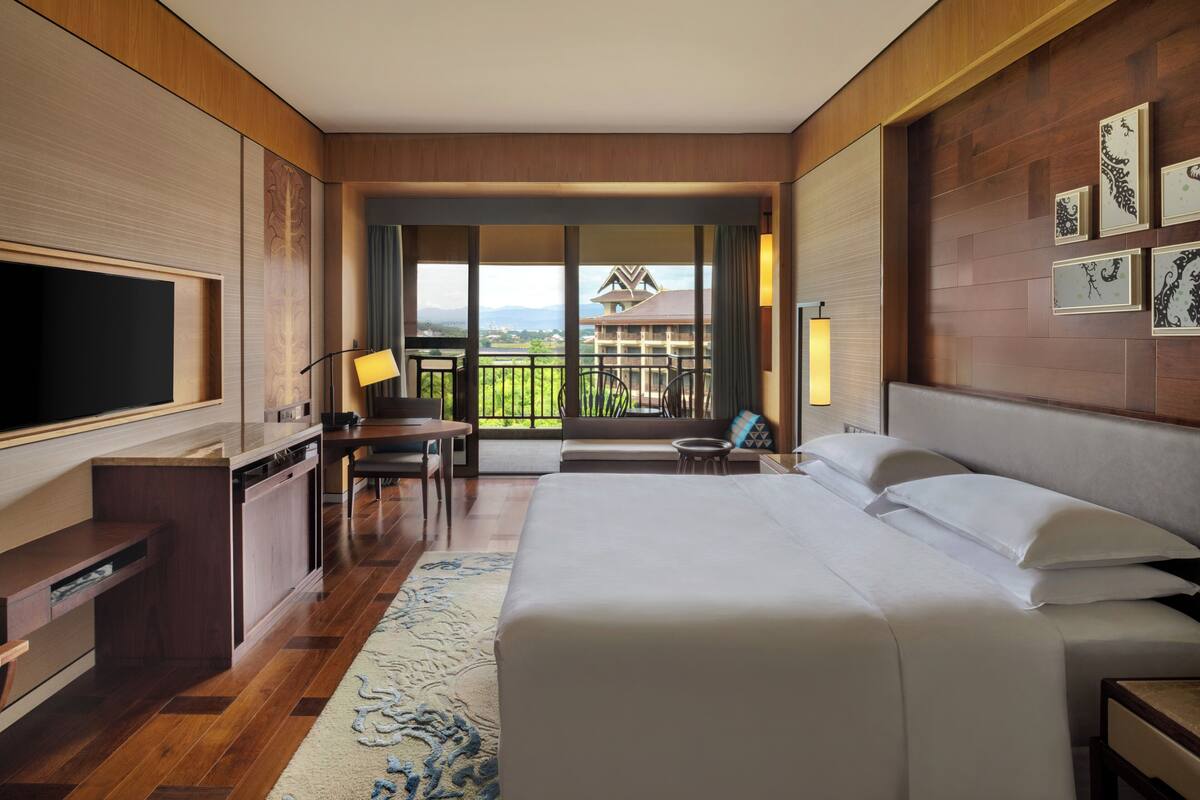 A hotel room with a large bed, a desk with a chair, a television, and a balcony with a view of buildings.