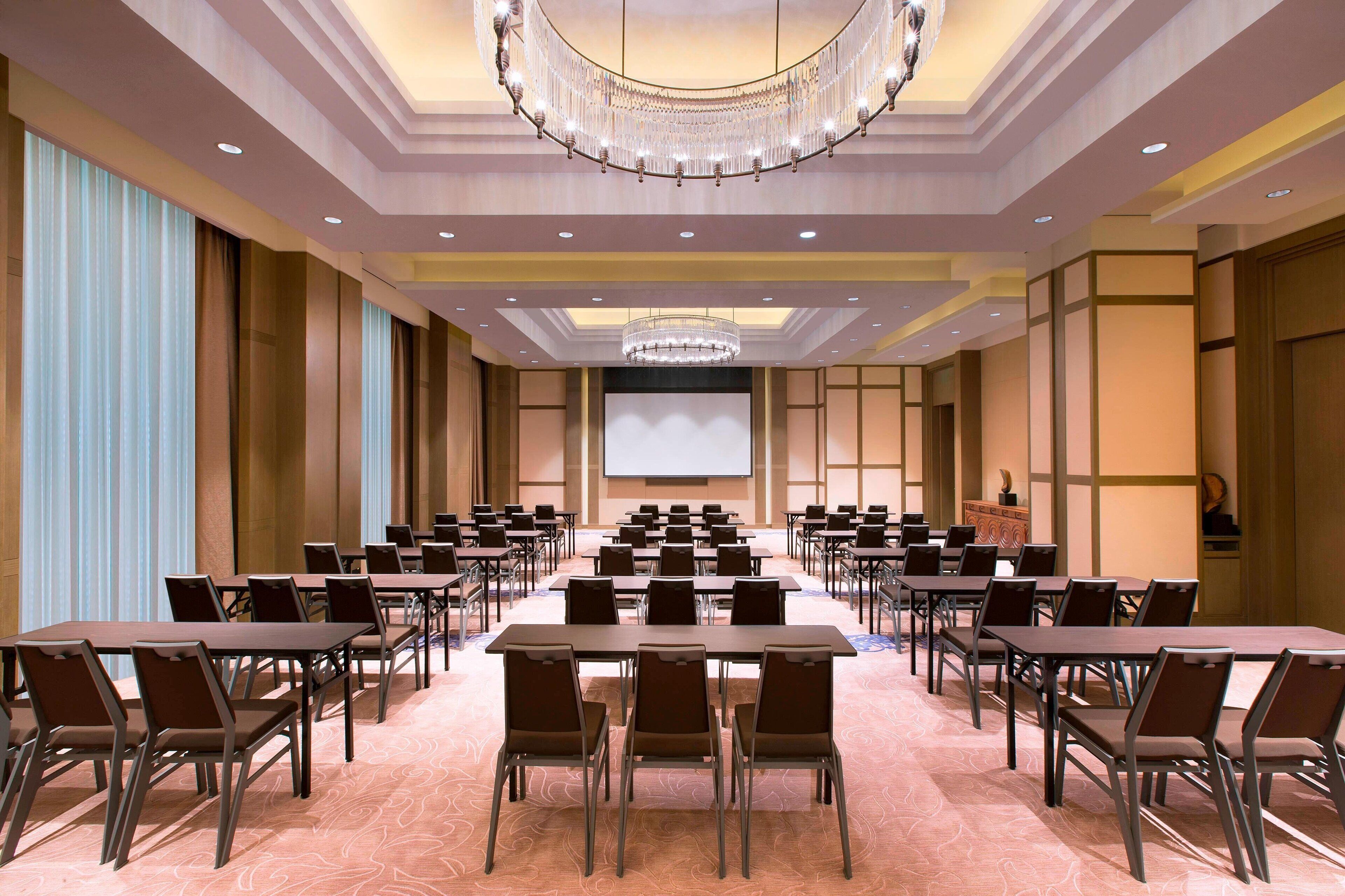 A conference room with rows of tables and chairs, a large screen at the front, and a chandelier.