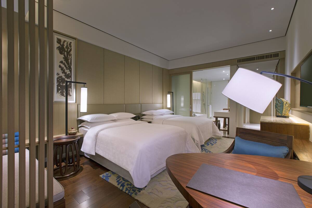 A hotel room with two beds, a desk, a chair, a lamp, and a large mirror.
