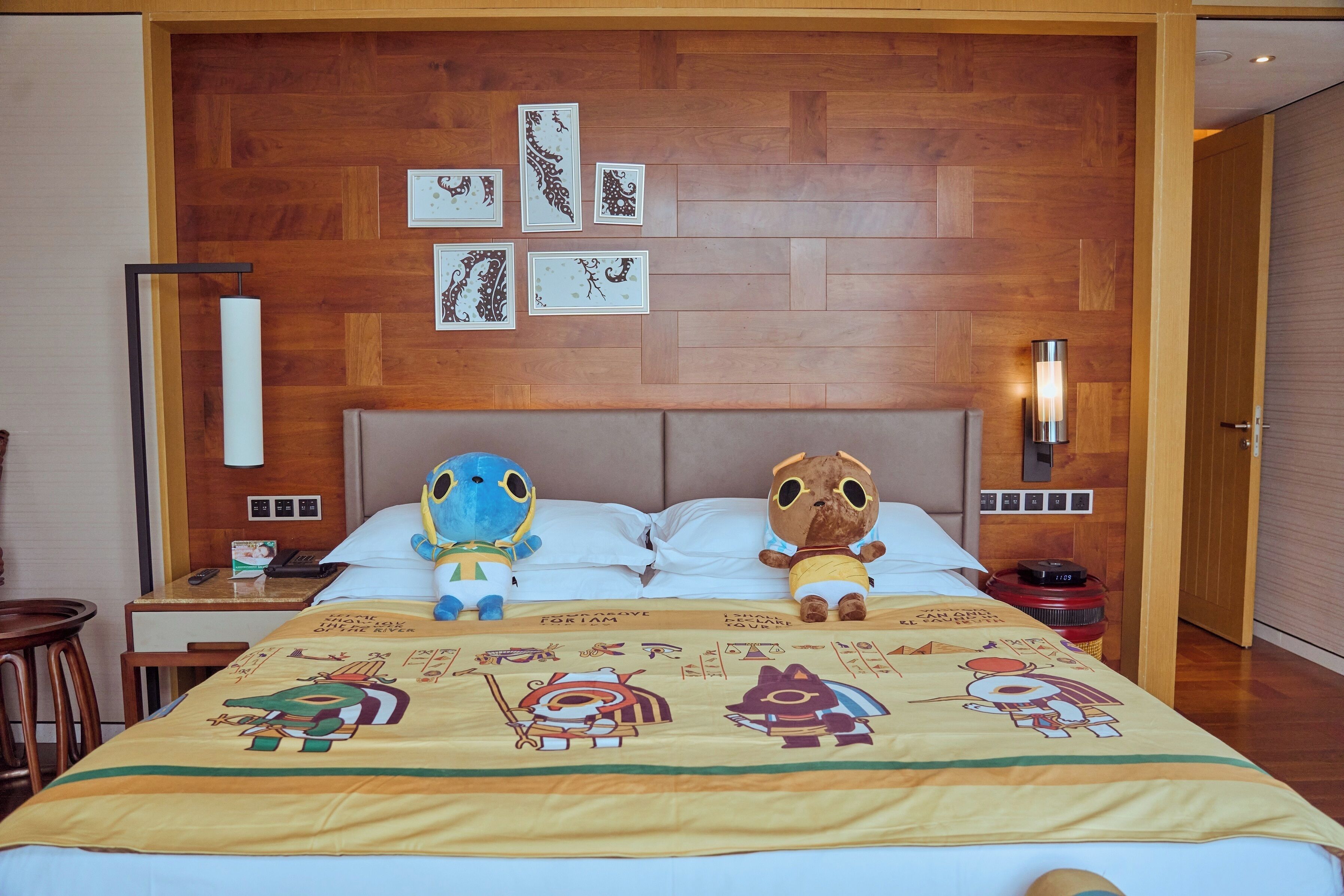 Une chambre d’hôtel avec un mur en bois, un lit recouvert d’une couette aux motifs de dessins animés, deux peluches, une table de chevet avec une lampe et un téléphone fixé au mur.