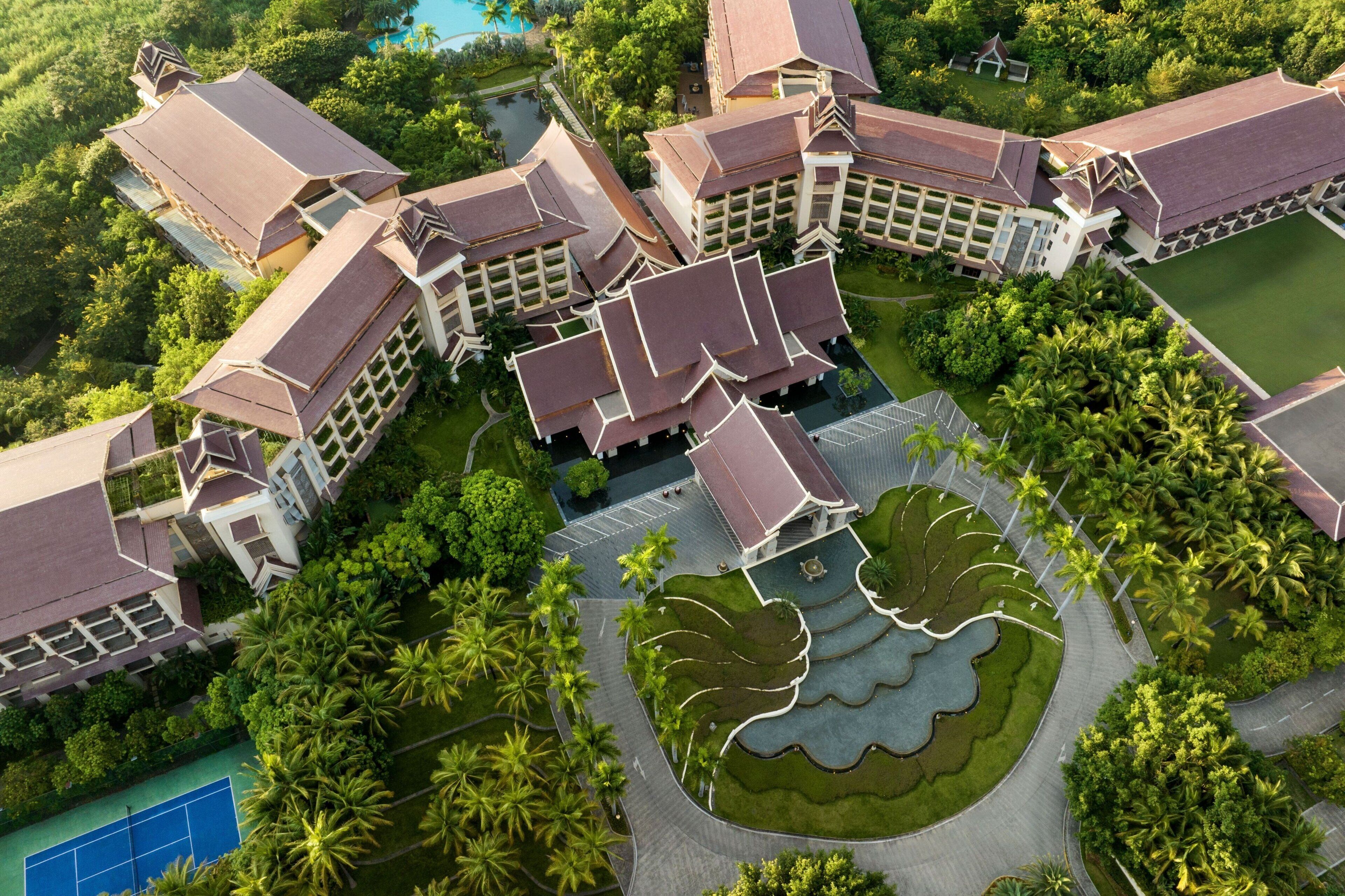 An aerial view of a resort with multiple buildings, a central courtyard with a water feature, and surrounding lush greenery.