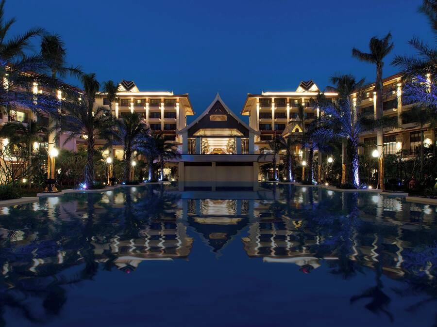 Um hotel com uma grande piscina espelhada em primeiro plano, rodeado por palmeiras e edifícios bem iluminados.