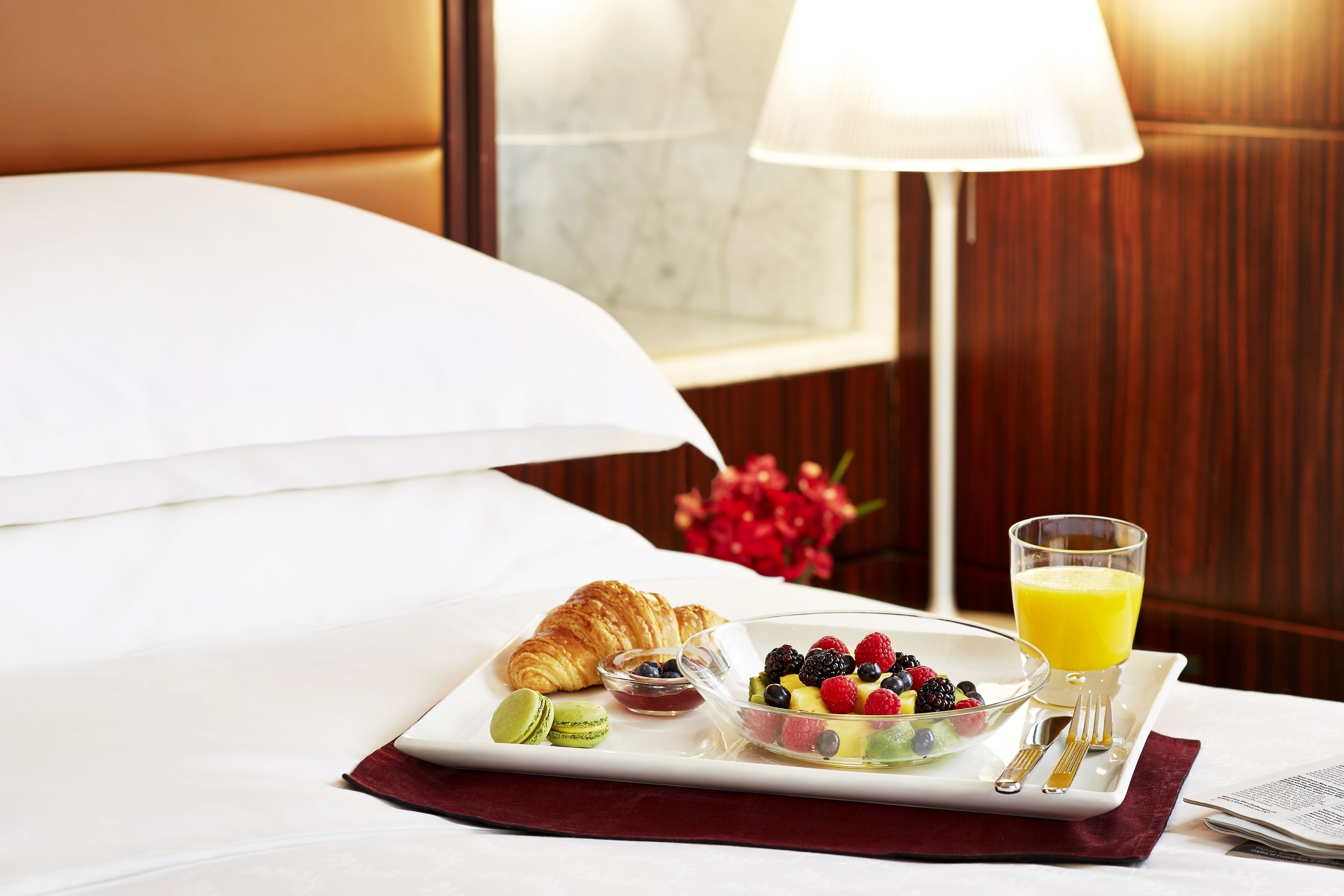 A breakfast setup on a hotel bed with croissants, a fruit bowl, and a glass of orange juice.