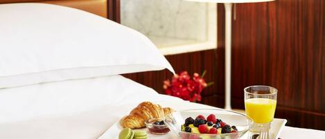 A breakfast setup on a hotel bed with croissants, a fruit bowl, and a glass of orange juice.